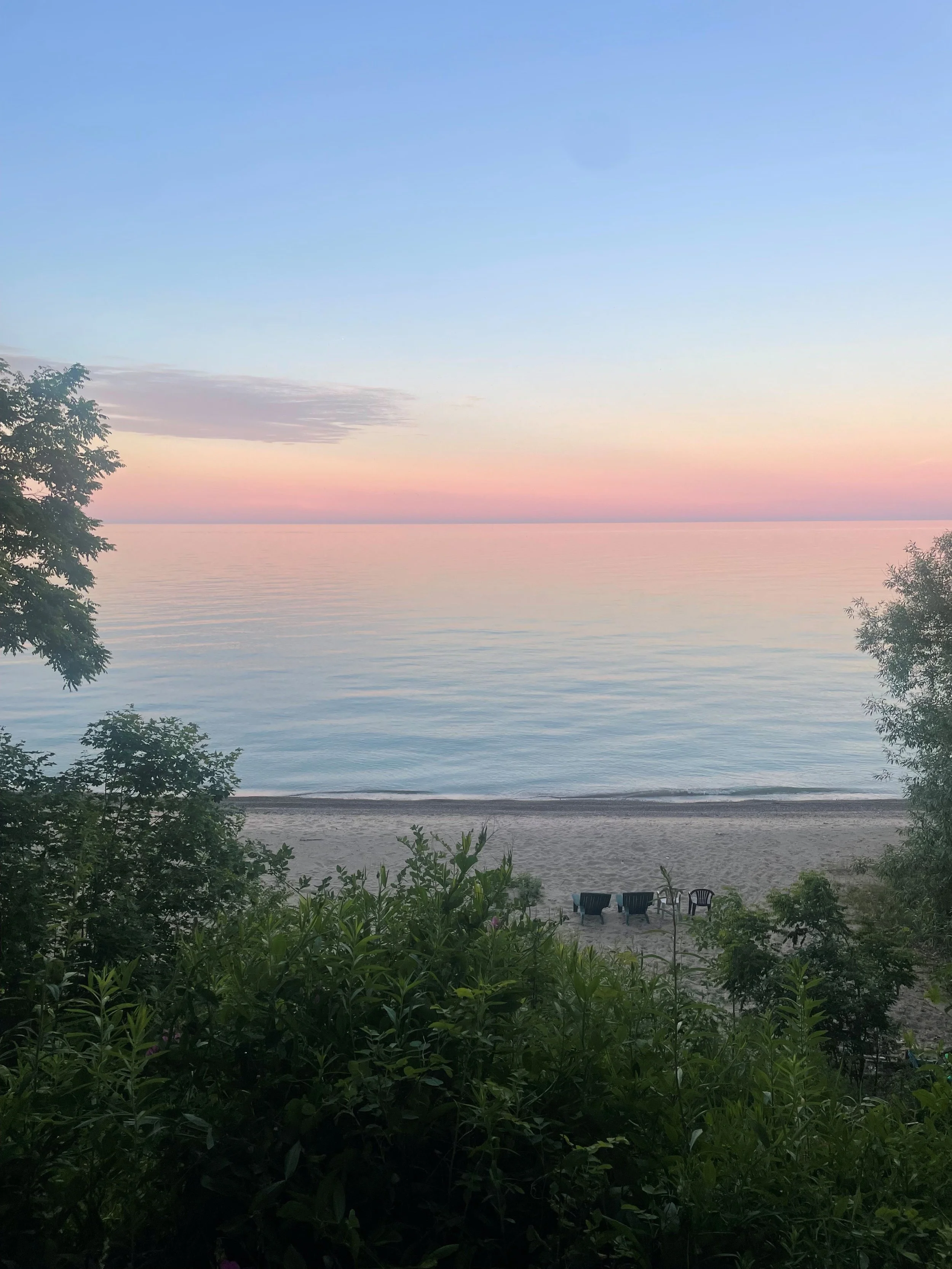 A peaceful sunset over a calm lake, with three chairs on the sandy beach and lush green bushes and trees in the foreground.