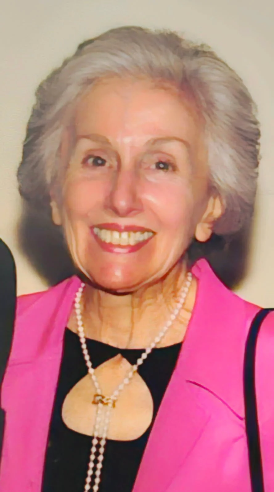 A smiling elderly woman with short gray hair wearing a pink blazer, black top with a keyhole neckline, a pearl necklace, and a pink lipstick.
