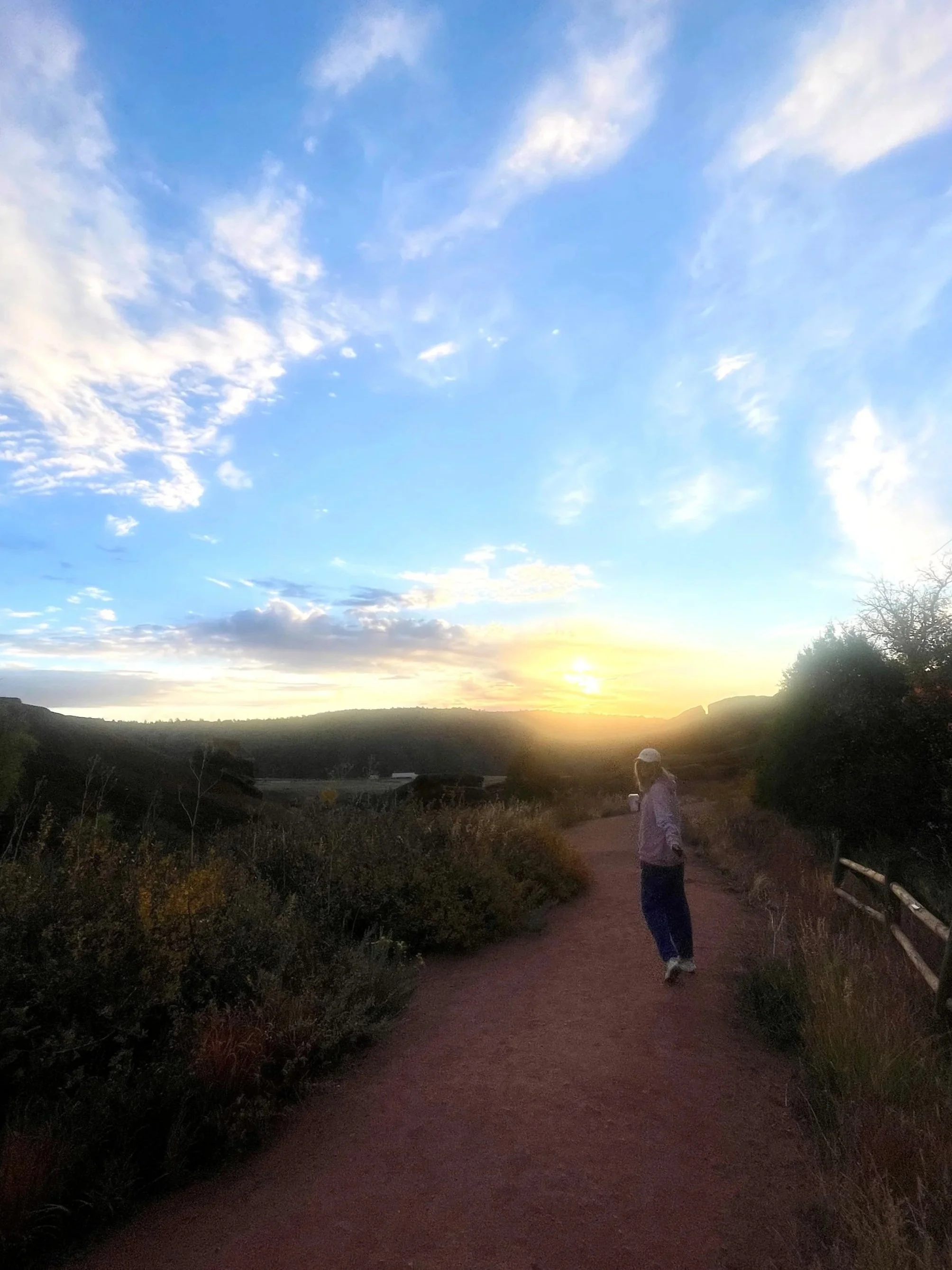 A person walking on a dirt path in a rural landscape during sunset with a partly cloudy sky.