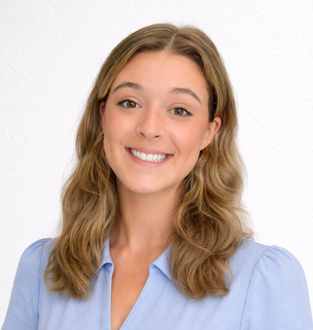 Portrait of a young woman with shoulder-length, wavy blonde hair, smiling, wearing a light blue collared shirt, against a white textured background.