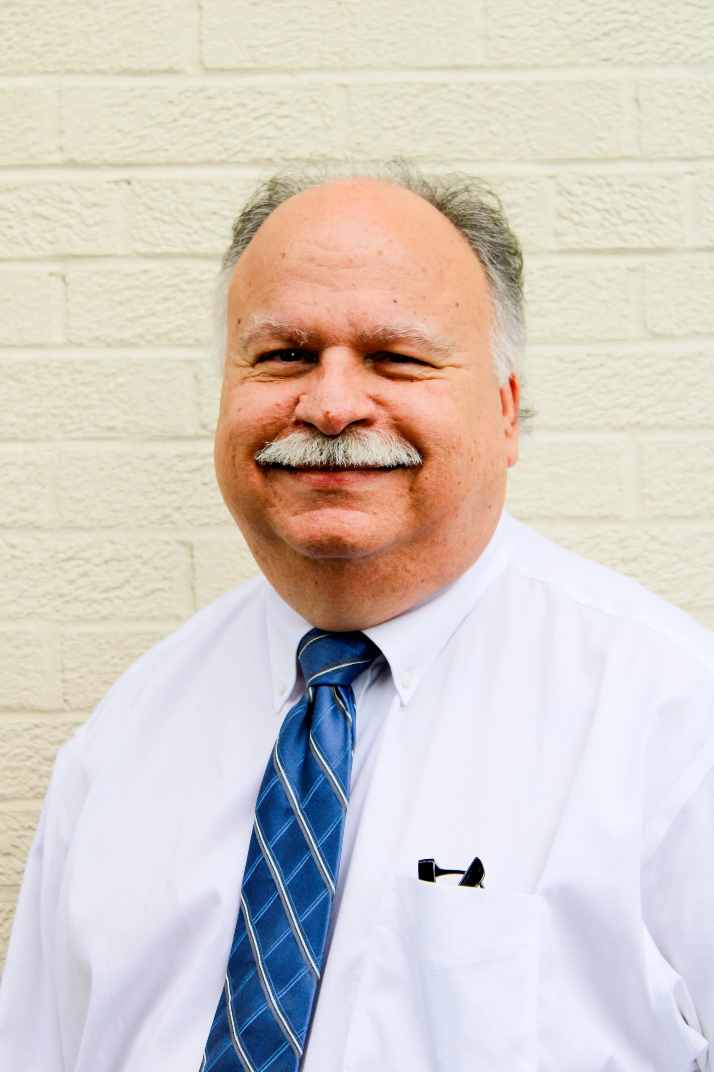 Portrait of a middle-aged man with a mustache, wearing a white shirt, blue tie, and glasses in his shirt pocket, standing in front of a beige brick wall.