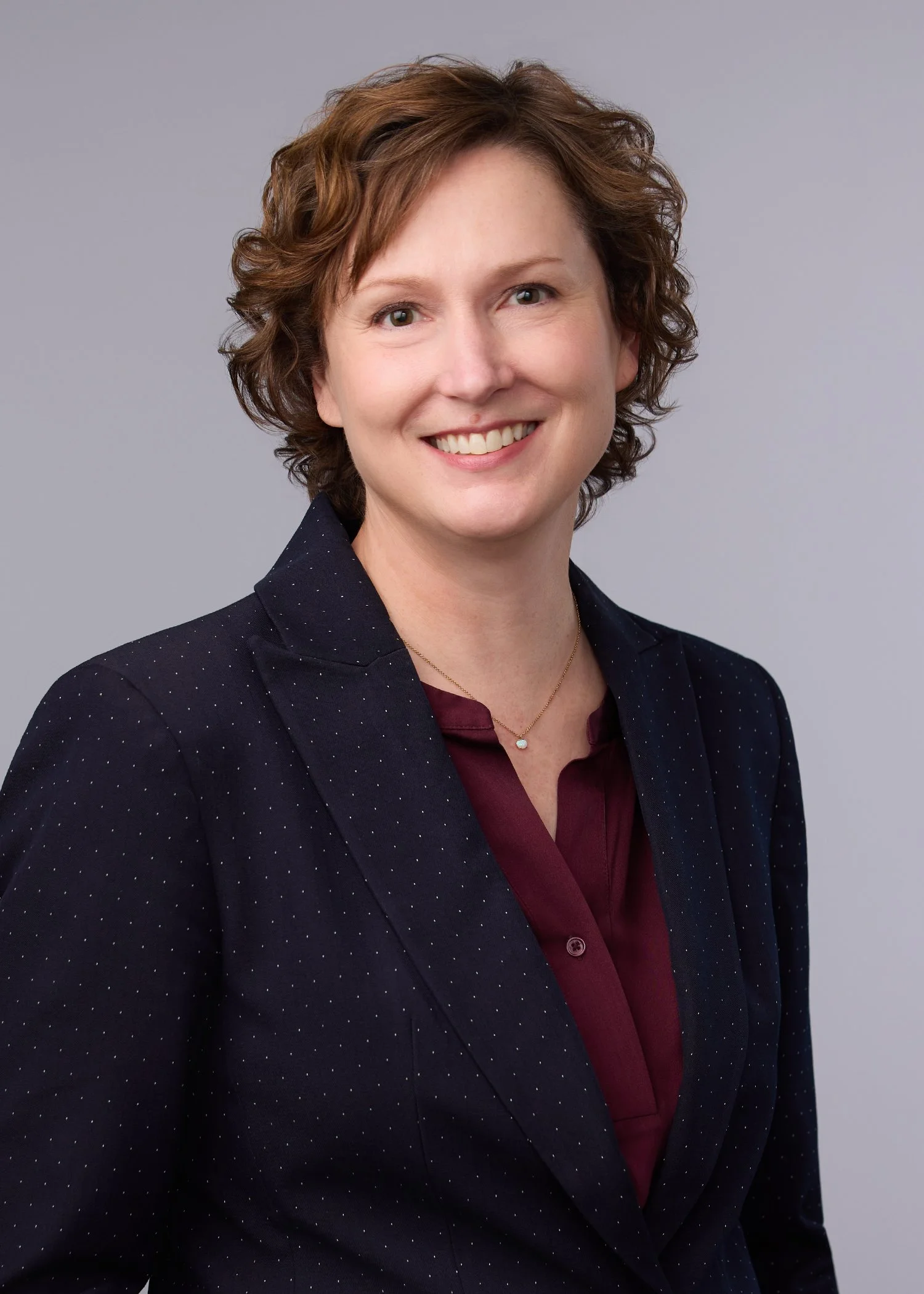 A professional headshot of a woman with short, curly, light brown hair, smiling, wearing a dark blazer with small white polka dots over a maroon blouse, and a small gold necklace, against a plain light gray background.