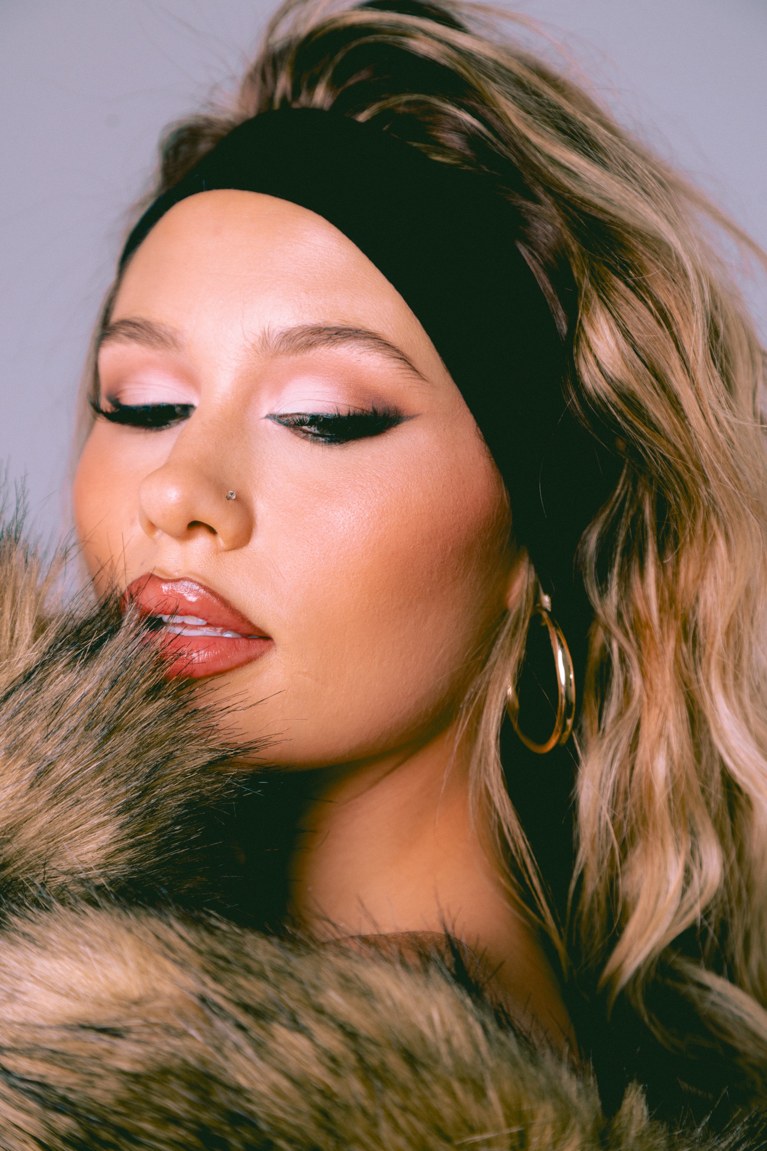 A close-up portrait of a young woman with light brown hair wearing a black headband, light-colored eyeshadow, black eyeliner, cut crease eye shadow and hoop earrings, partly covered by a fur coat.