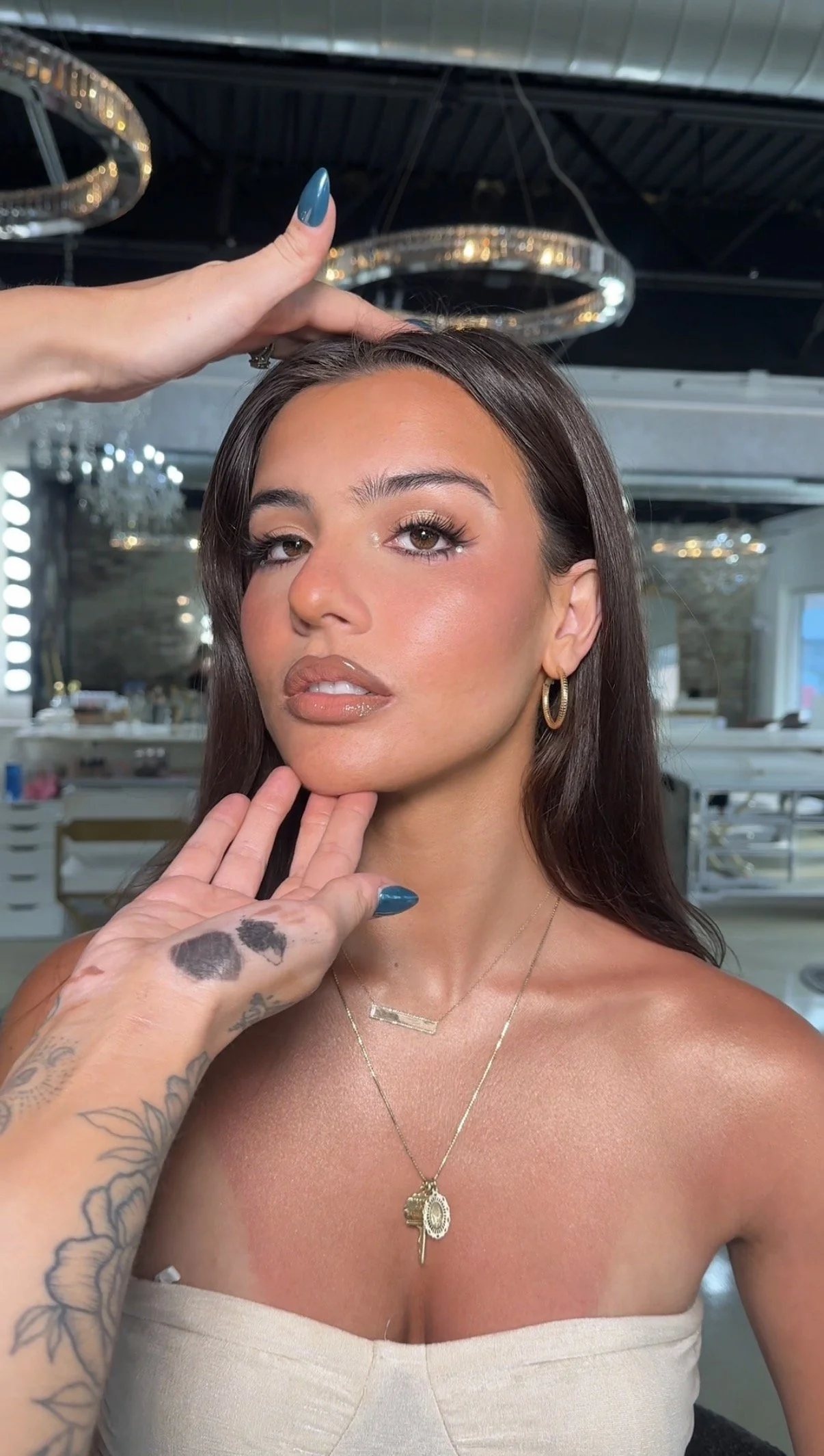 A woman with long dark hair, wearing gold hoop earrings and layered necklaces, has makeup on and an earring in her left ear. A person's tattooed hand with blue painted nails is touching her face and head. The background features elegant chandeliers and a modern interior.