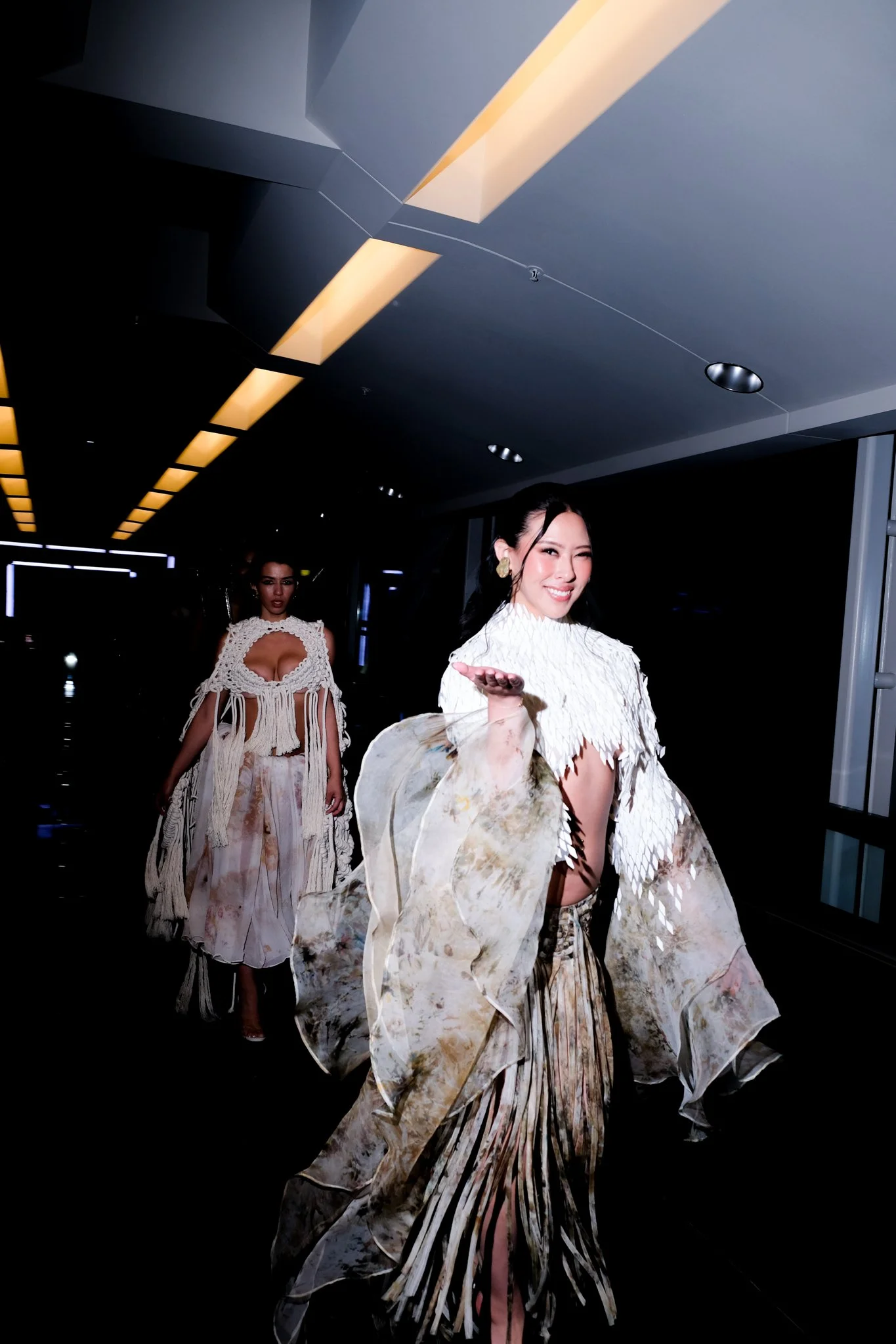 Two women in modern fashion walking in a fashion show. They wear elaborate, cream-colored outfits with textured fabrics and flowing fabrics, and are smiling and posing. They have professional, well done makeup on.