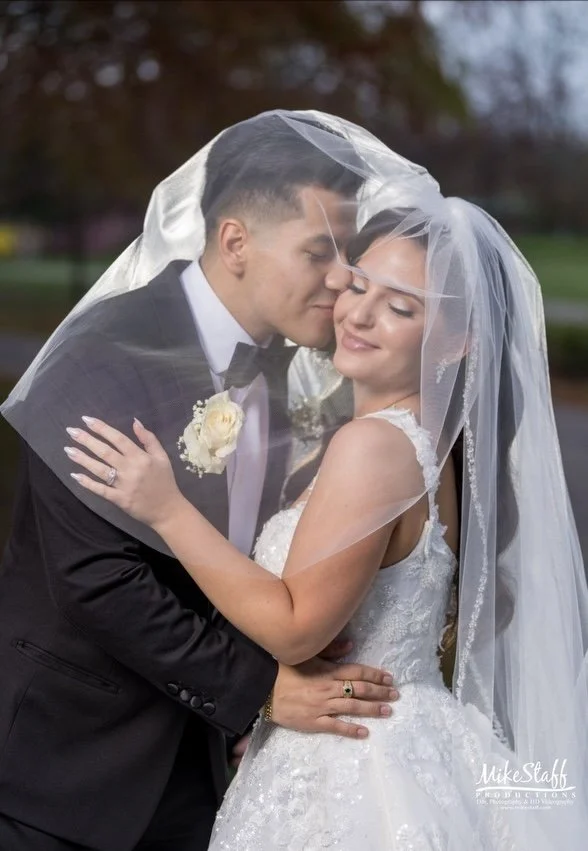 Bride and groom embracing outdoors, dressed in wedding attire, under a veil, with trees and a park in the background. Bride has glowing skin, soft glam makeup, pink lips