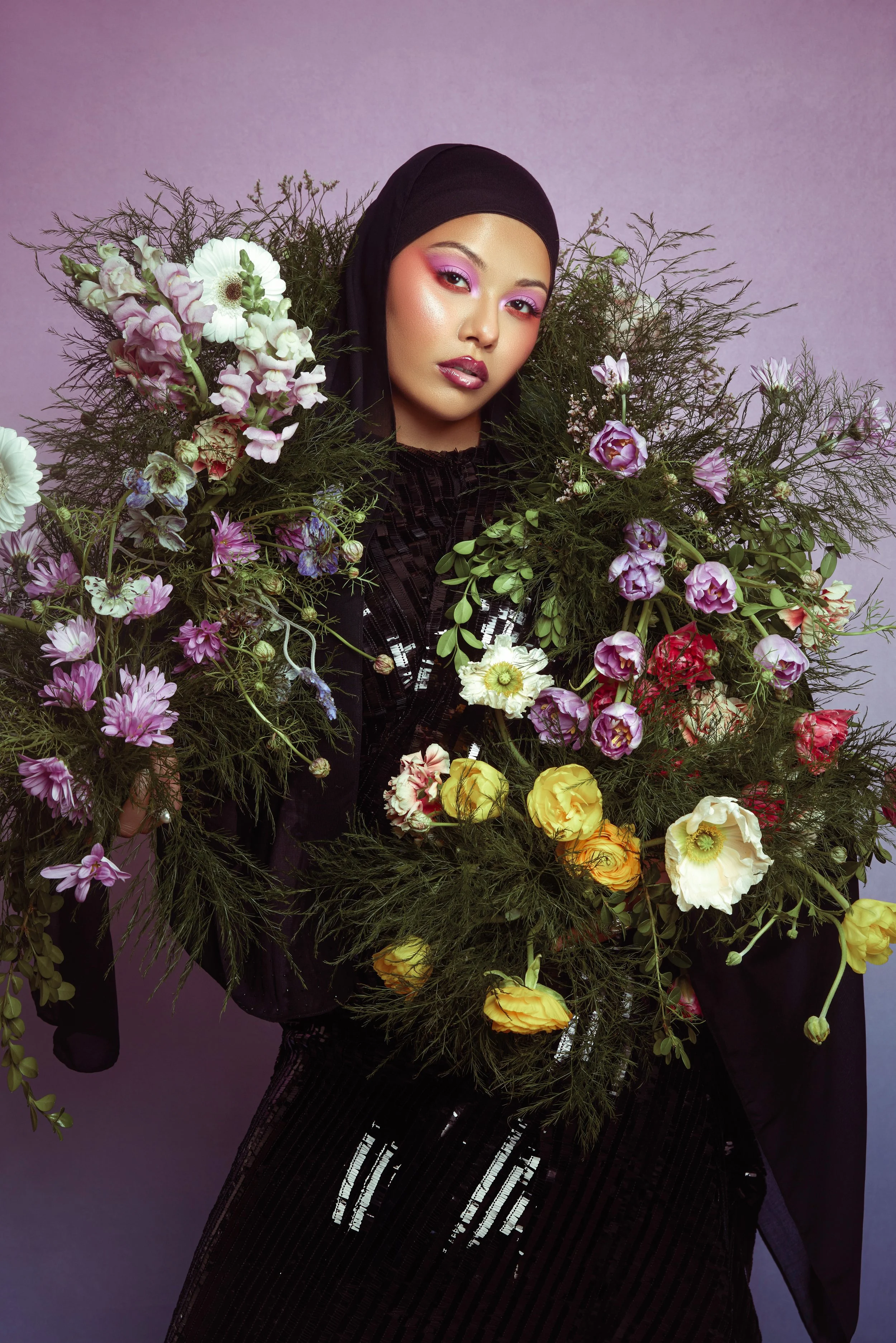 A woman wearing a black hijab, with bold pink eyeshadow, standing against a purple background, holding a large bouquet of mixed pastel and brightly colored flowers.