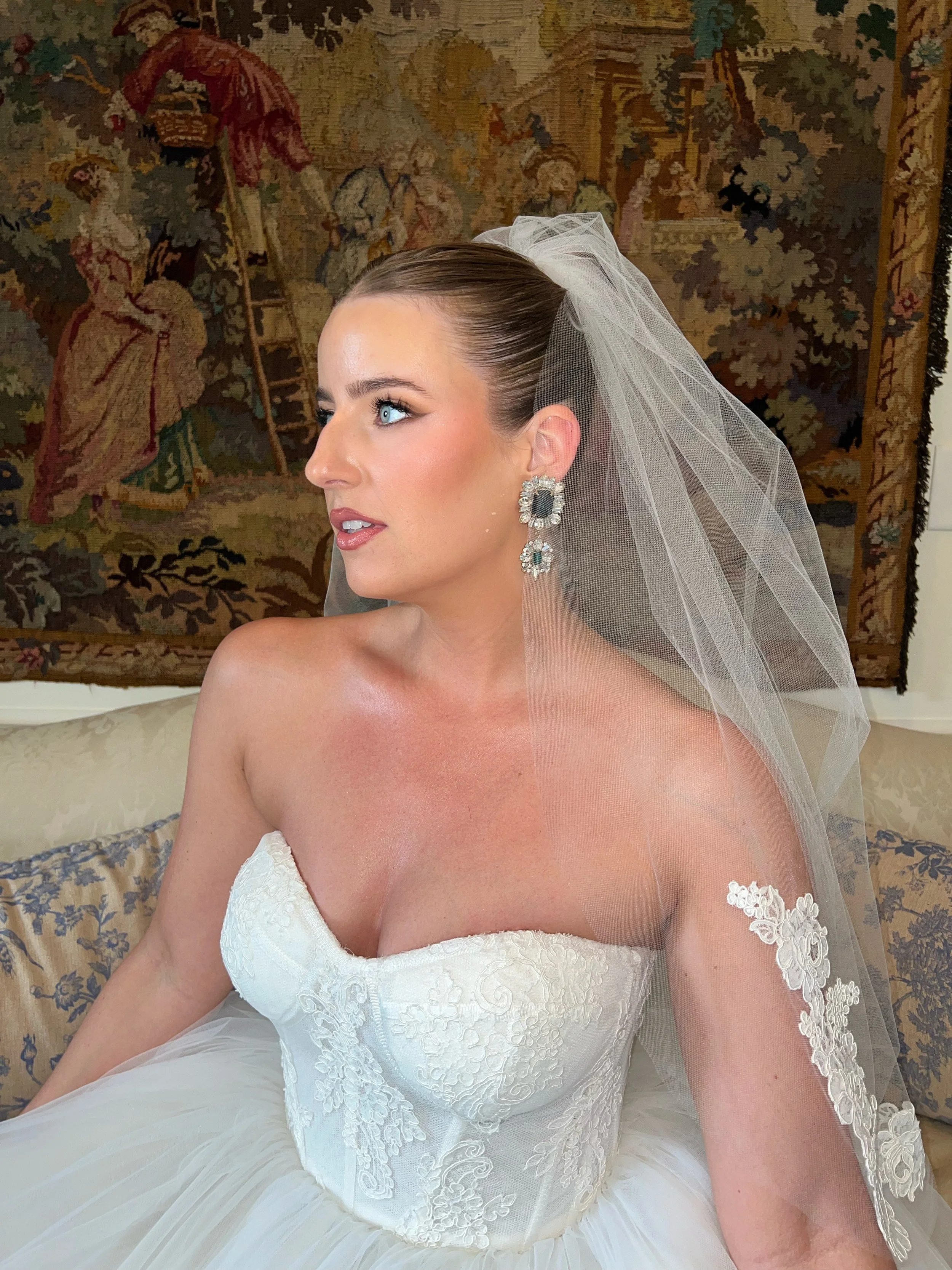 A bride with blue eyes wearing a white lace wedding dress, a tulle veil, and large earrings, sitting in front of a tapestry with a scene of people, trees, and a ladder.