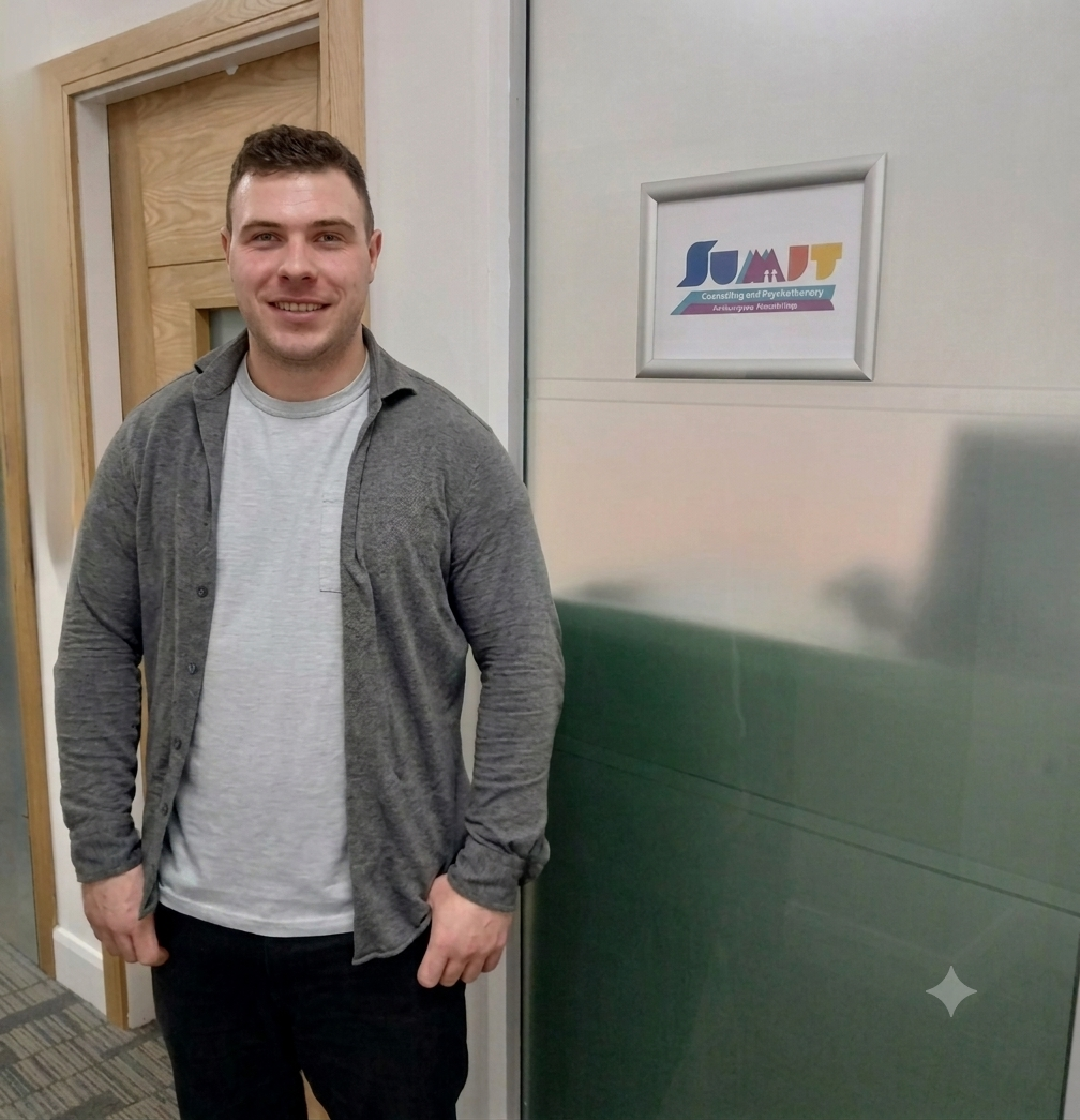 A smiling young man with short brown hair wearing a gray cardigan over a light gray t-shirt, standing indoors next to a wall with a framed sign. The sign has colorful text and a logo, and the background includes a wooden door.