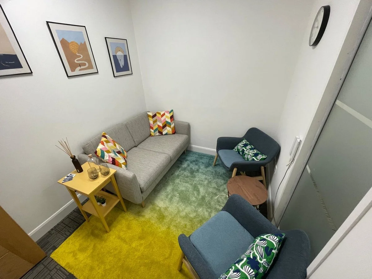 Small living room with a gray sofa, two patterned throw pillows, a yellow side table with decorative items, a green and yellow rug, two blue armchairs with floral pillows, a small wooden side table, framed art on the wall, and a clock.