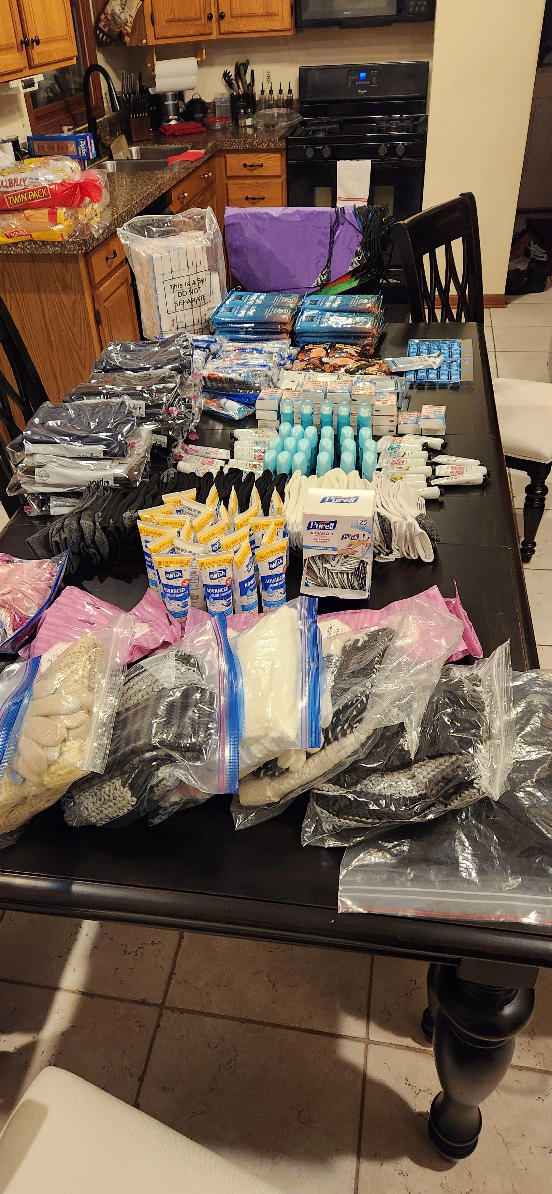Table filled with various items, including socks, hygiene products, and supplies, in a kitchen with wooden cabinets and stainless steel appliances.