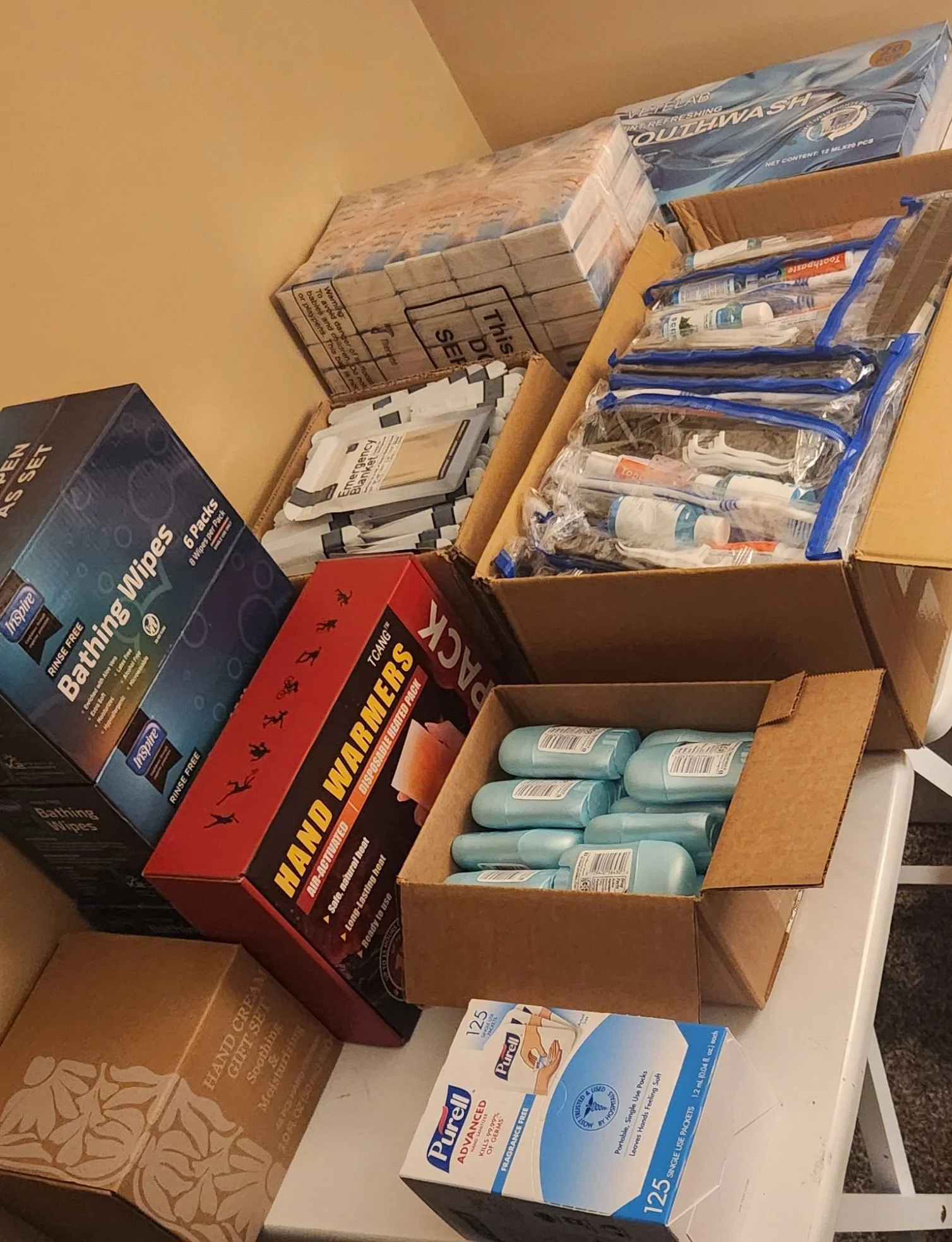 Various household supplies including boxes of disposable bathroom wipes, boxes of face masks, a large box of hand and body sanitizer bottles, and a box of large pack of facial tissues, arranged on a white table and cardboard boxes.