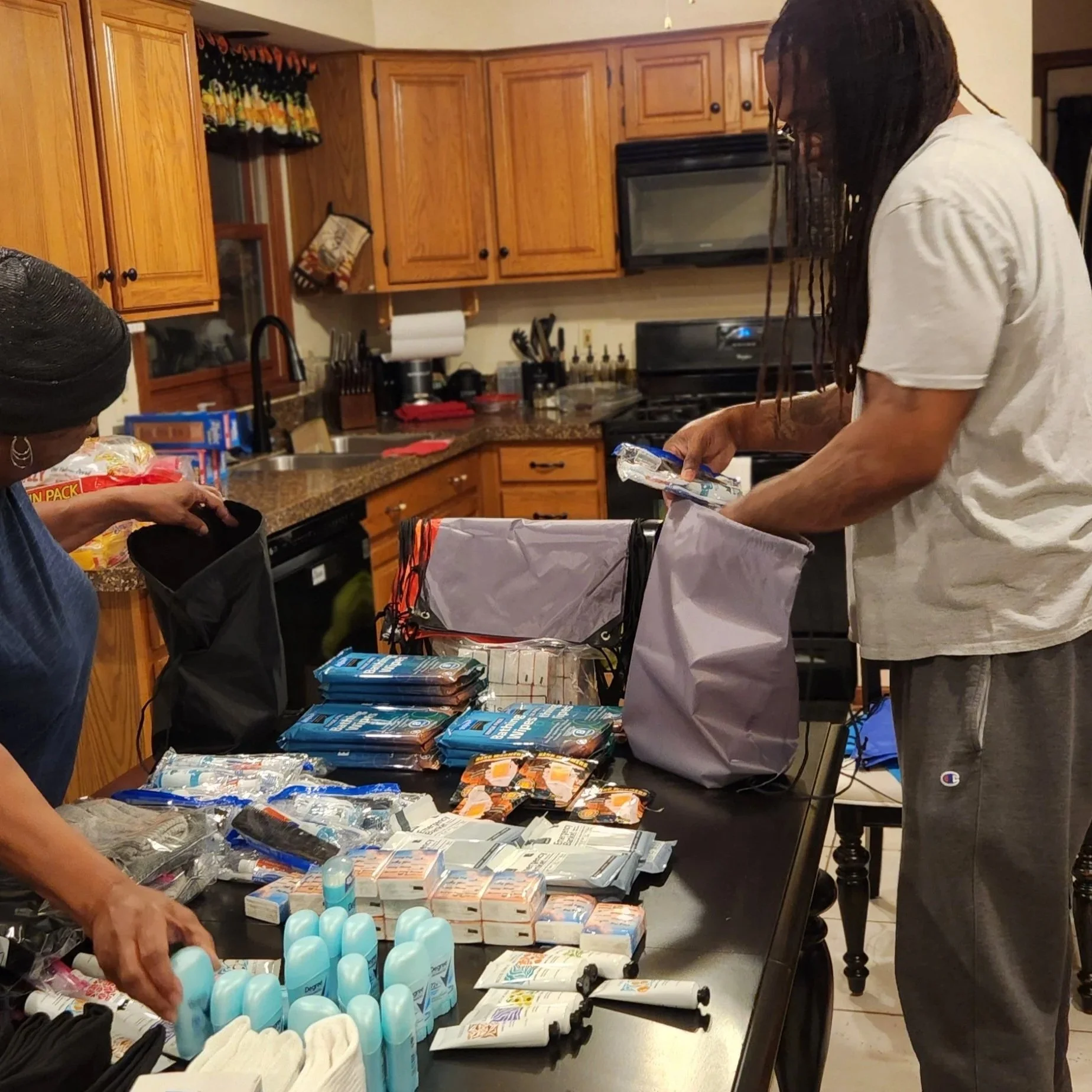 A woman with braided hair and glasses in a white T-shirt sorting personal care items on a kitchen table, with packaged products including lotions, shampoos, and hygiene items, in a kitchen with wooden cabinets and black countertops.