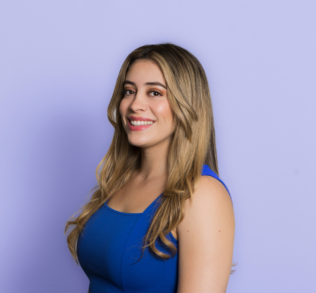 Young woman with long blonde wavy hair smiling in a blue sleeveless top, standing against a lavender background.
