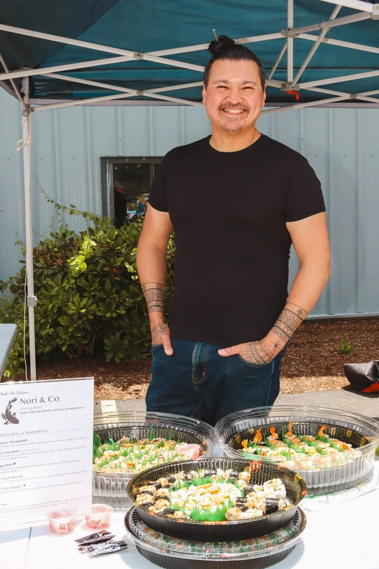 A man, Chef Jayson, is wearing a black t-shirt, smiling while standing behind a display of various sushi trays at an outdoor event. There is a sign for Nori & Co on the table and people visible in the background.