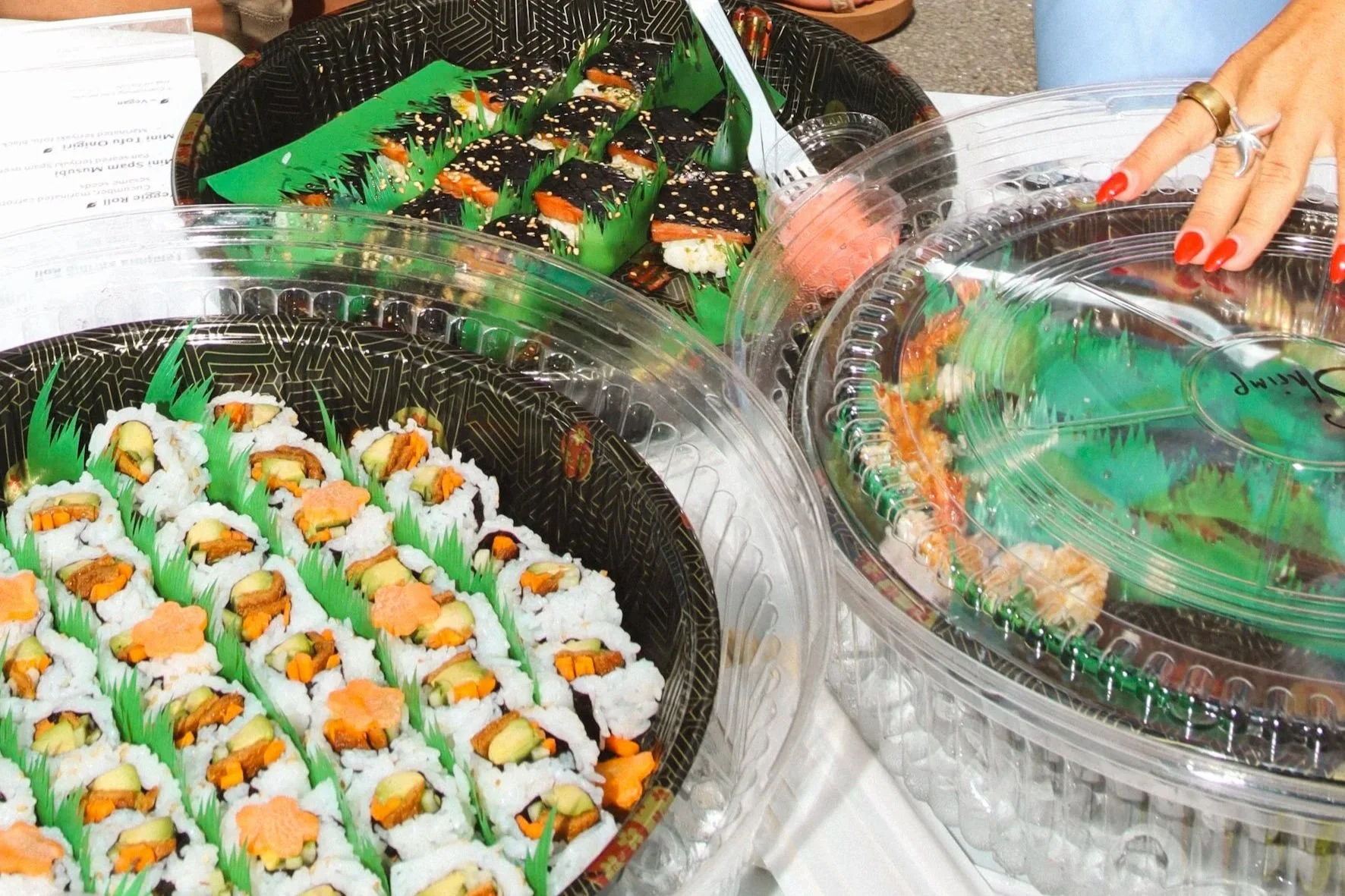 Sushi in black snack boxes with green divider inserts, including sushi rolls with fish and vegetables, and sushi pieces with black seaweed and sesame seeds, being stored or displayed in clear plastic containers.