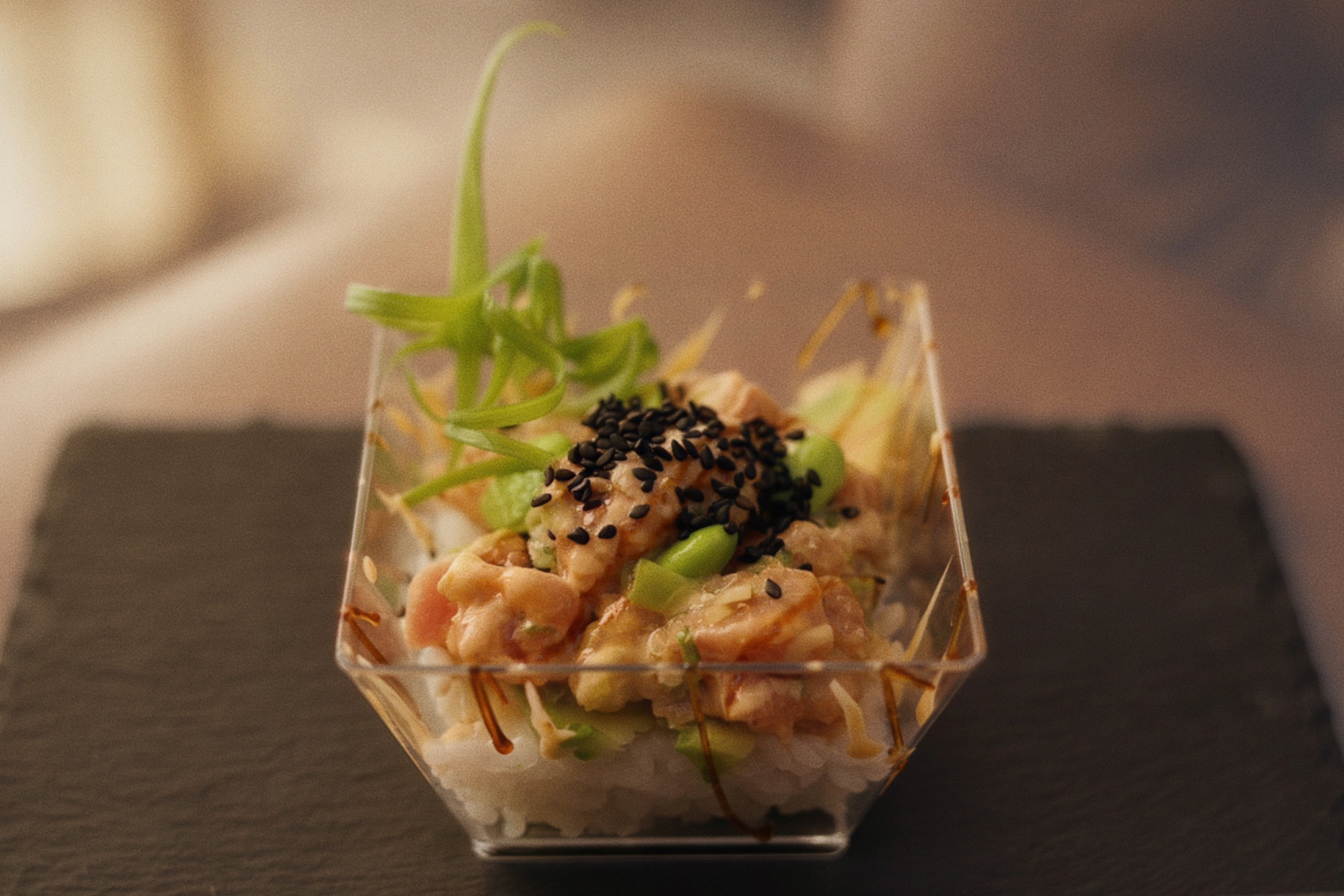 A close-up view of a small appetizer or dish served in a clear, geometric glass container. It contains chopped seafood or chicken, topped with black sesame seeds, green sauce, and garnished with microgreens or herbs. The dish is placed on a dark surface with a blurred background.