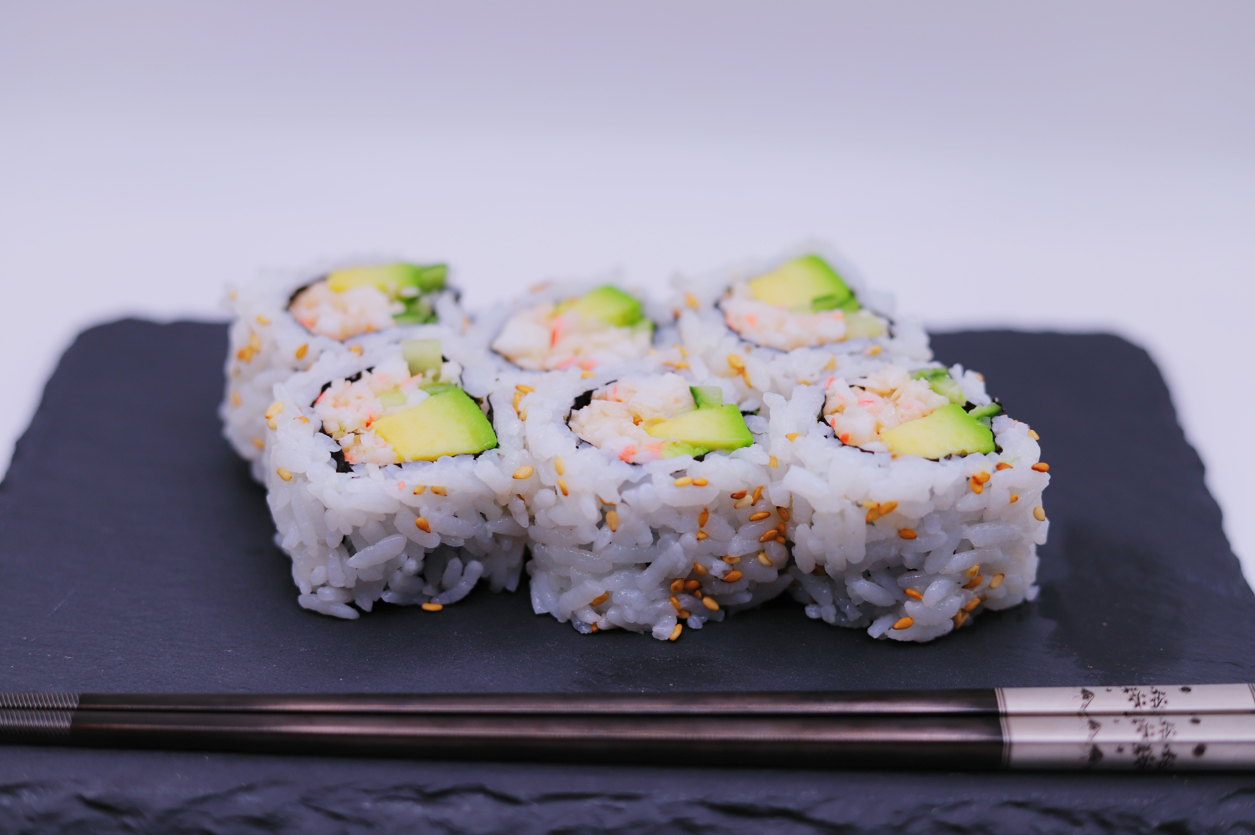 Close-up of sushi rolls on a black slate serving board with chopsticks underneath, featuring rice, avocado, crab meat, and sesame seeds.