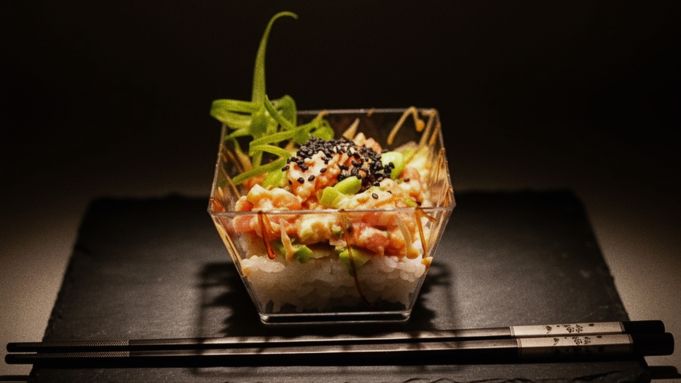 A gourmet dish served in a small glass bowl with rice, chopped vegetables, black sesame seeds, and a sprig of green garnish.