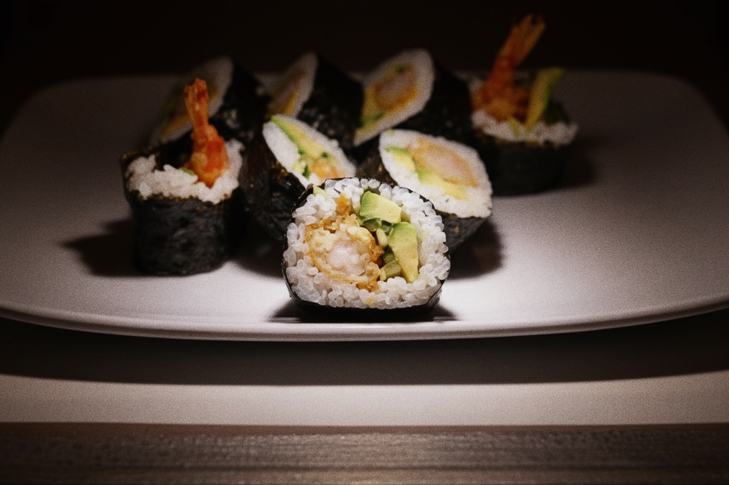 Close-up of sliced sushi rolls with shrimp, avocado, and rice on a white plate, dark background.