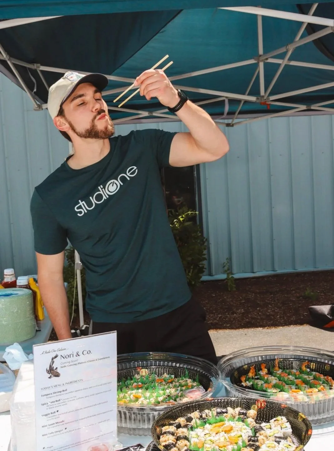 A man wearing a black T-shirt with the word 'studiolone' and a beige baseball cap is standing under a blue canopy, eating sushi with chopsticks. There are several trays of sushi on the table in front of him, along with a menu sign, condiments, and paper towels.