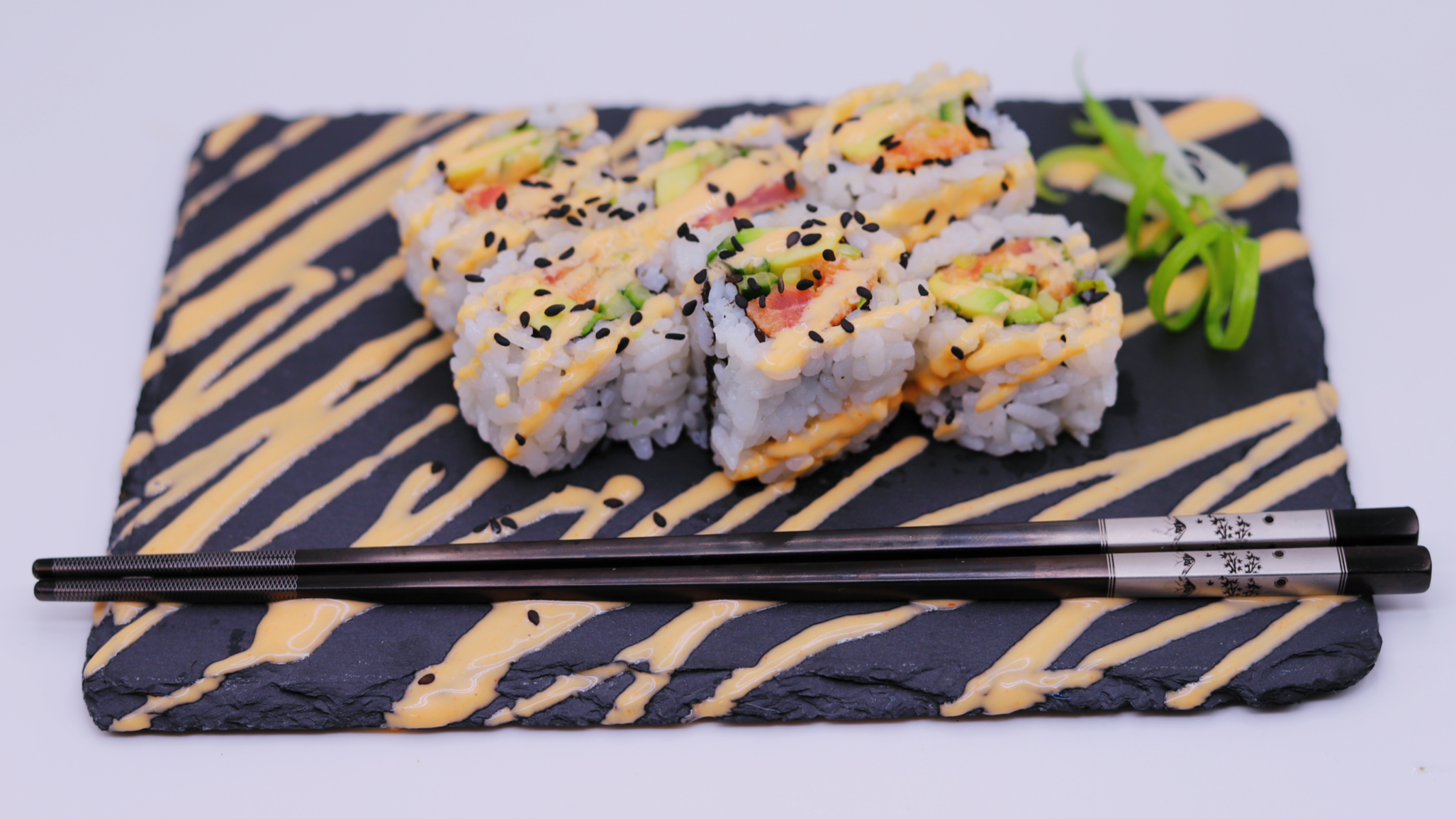 Sushi rolls topped with drizzle of sauce, black sesame seeds, and garnished with green onion on a black slate serving platter.