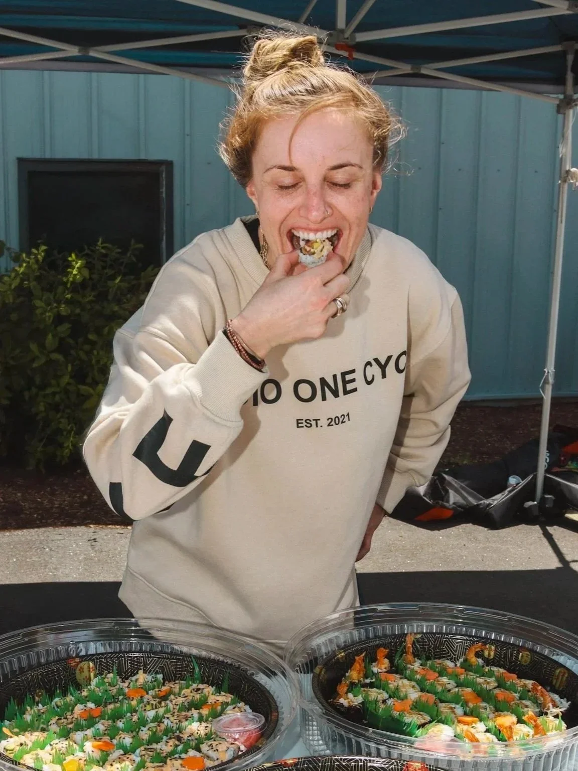 A young woman with reddish hair in a ponytail eating sushi from a tray at outdoor event.