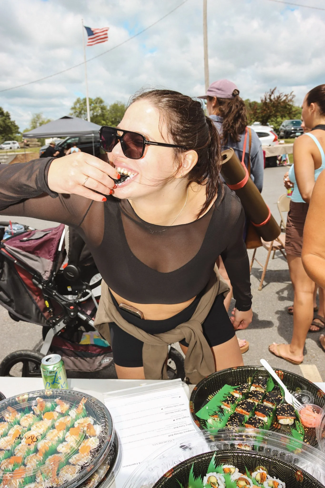 A woman with dark hair, wearing sunglasses and a black sheer top, is smiling while eating sushi at an outdoor event with other people. There are sushi platters on the table and an American flag in the background.