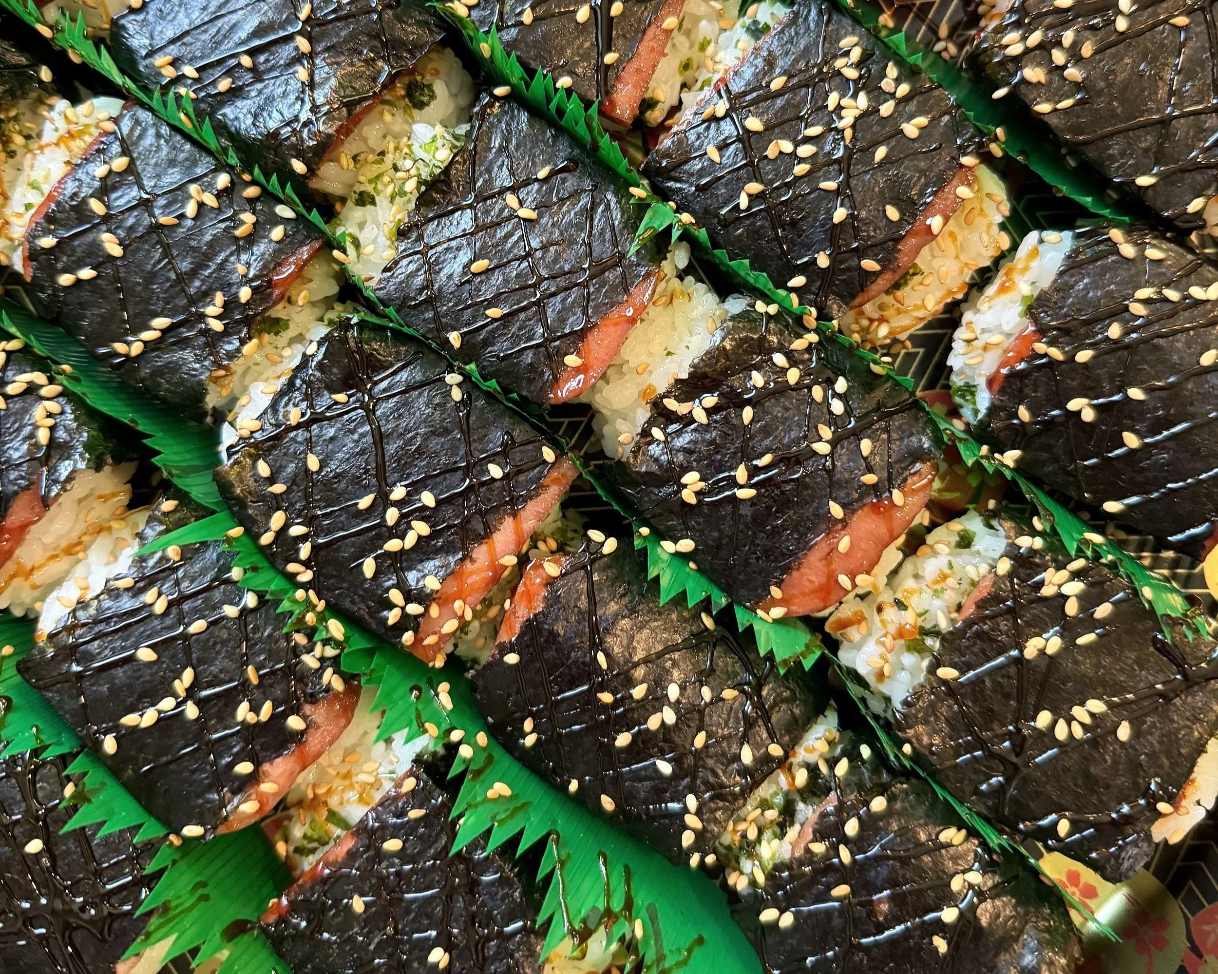 Close-up of sushi rolls wrapped in seaweed, topped with sesame seeds, drizzled with sauce, arranged on green trays.