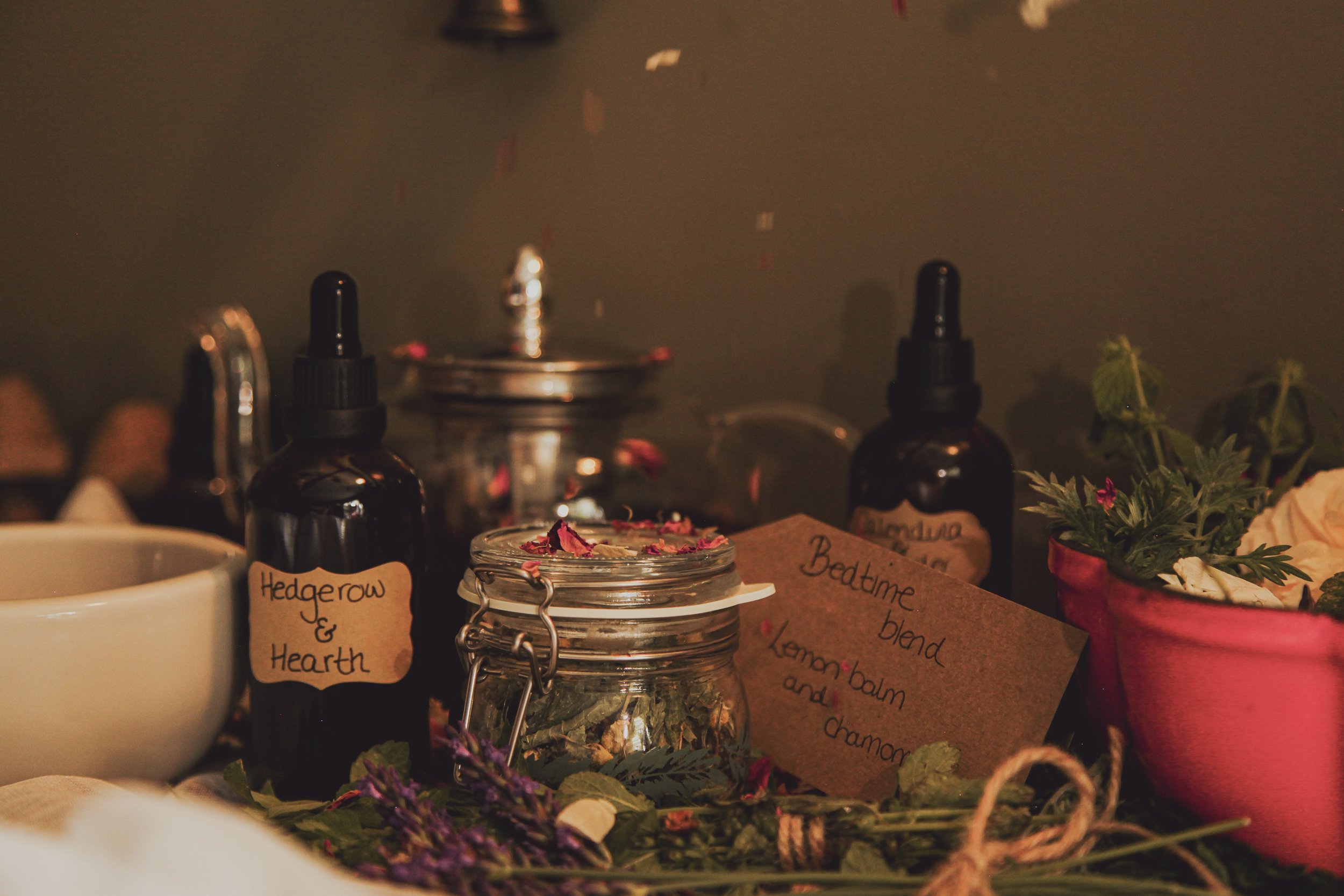 Close-up of a table with various bottles, jars, and handwritten labels. There are herbs and flowers around the items, and the background features a countertop with more bottles and a sink.