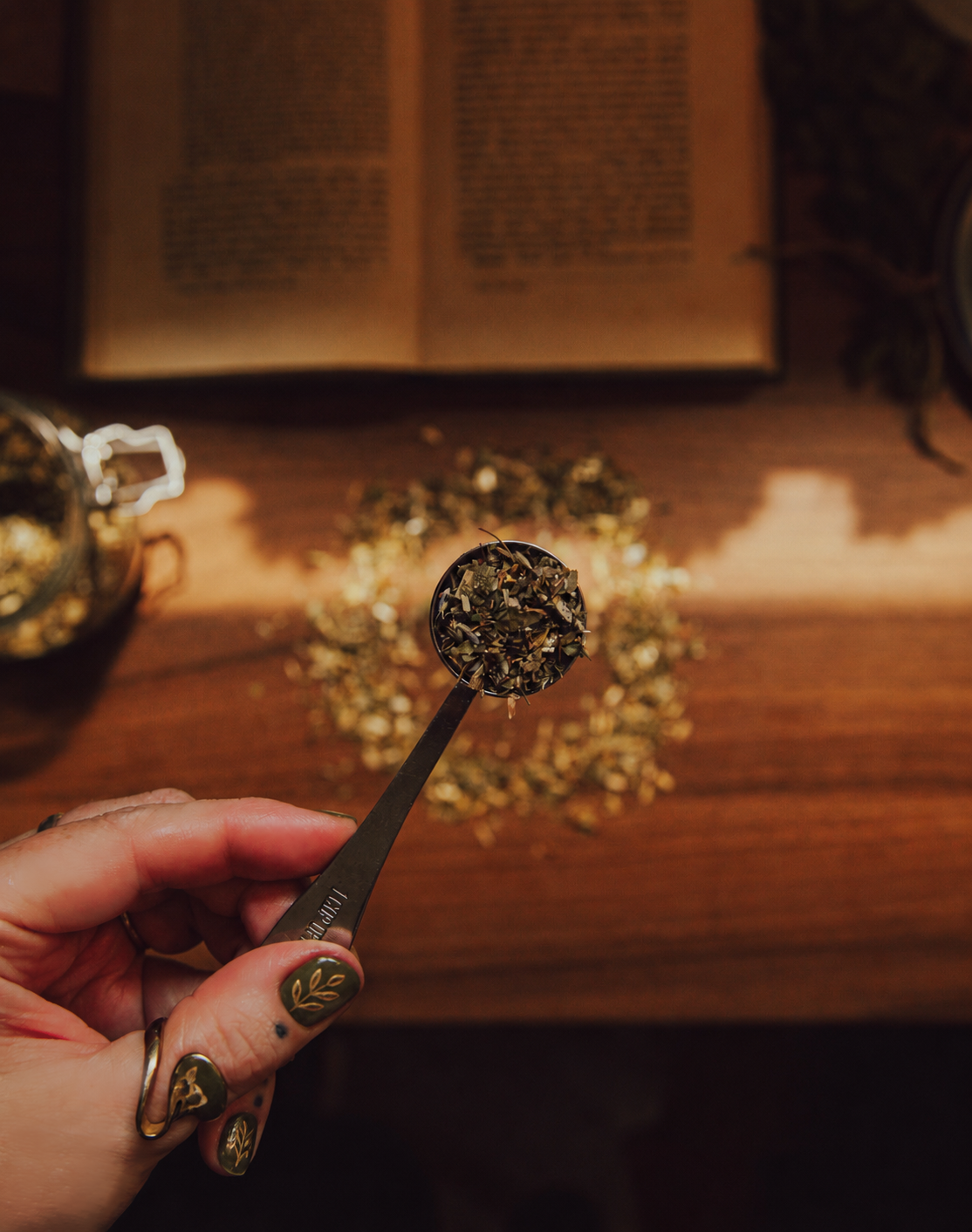 Close-up of a hand holding a metal spoon filled with dried herbs, with loose herbs scattered on a wooden table, an open book in the background, and a jar of herbs to the side.