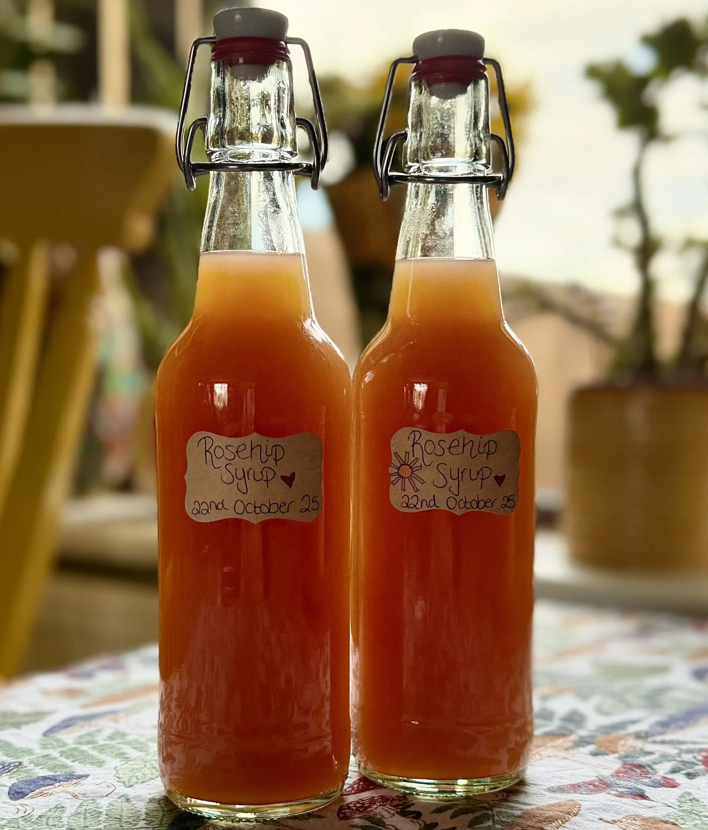Two glass bottles filled with orange-colored rosehip syrup, labeled with handwritten notes date October 22, 2025, on a floral tablecloth with blurred indoor background.