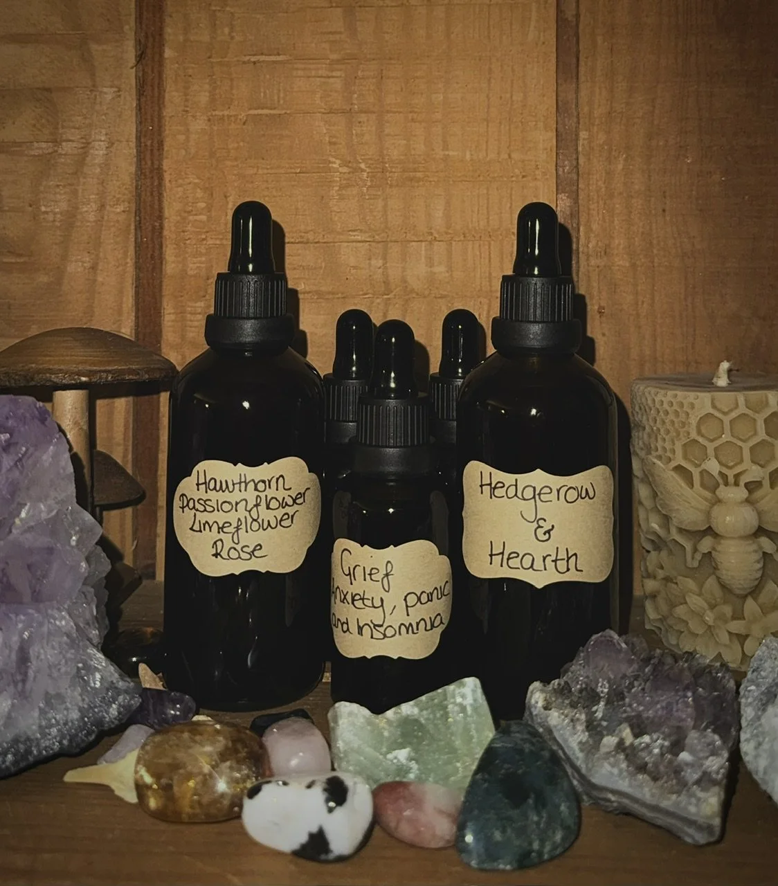 Dark glass dropper bottles labeled with handwritten notes that read 'Hawthorn Passionflower Lavender Rose,' 'Grief, Anxiety, Panic and Insomnia,' and 'Hedgerow & Earth,' surrounded by various crystals and stones, with a wooden wall background.