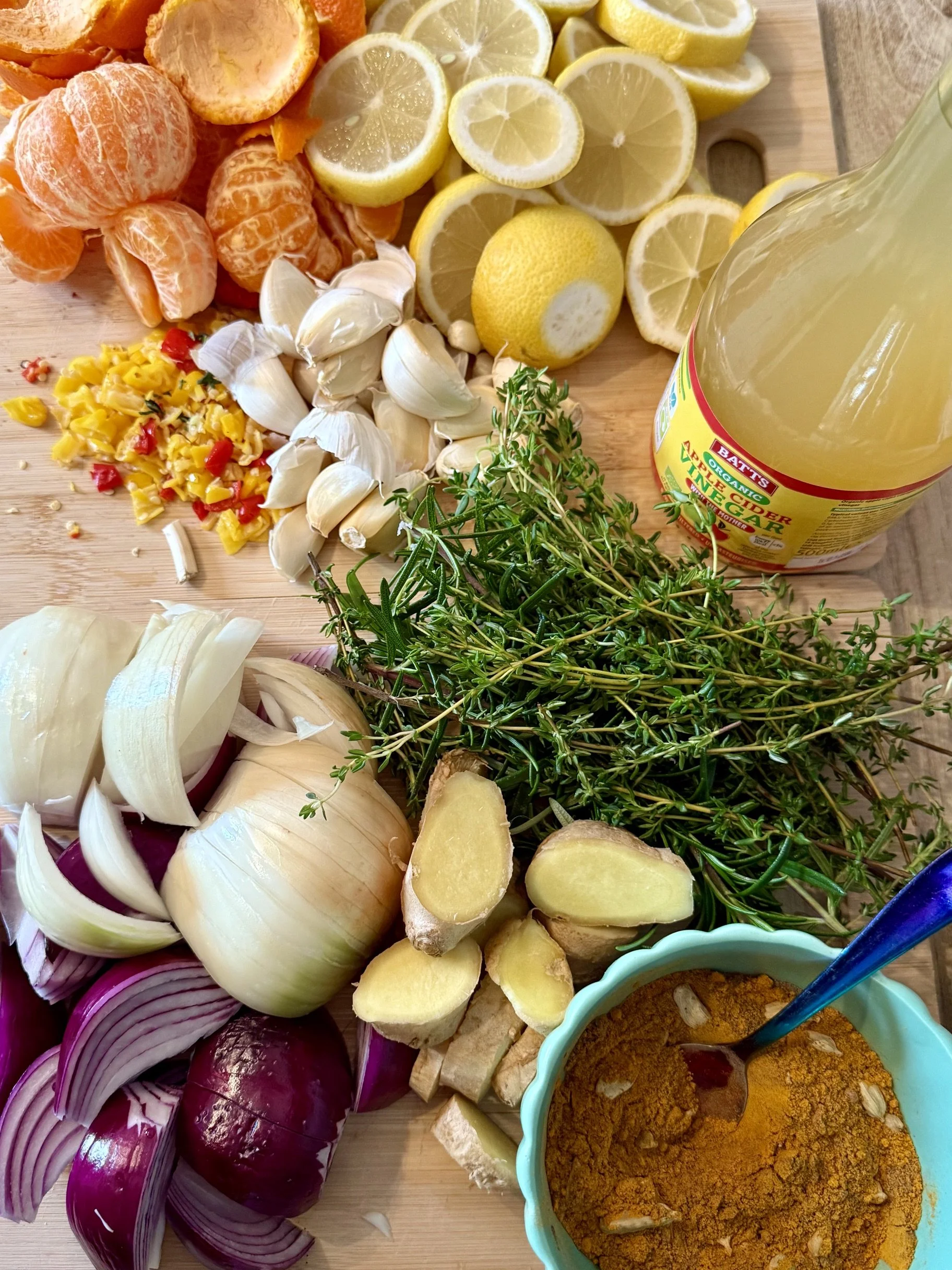 A variety of fresh ingredients for cooking or making curry, including onions, garlic, ginger, lemon, mandarin oranges, herbs, spices, and a bottle of apple cider vinegar on a wooden cutting board.