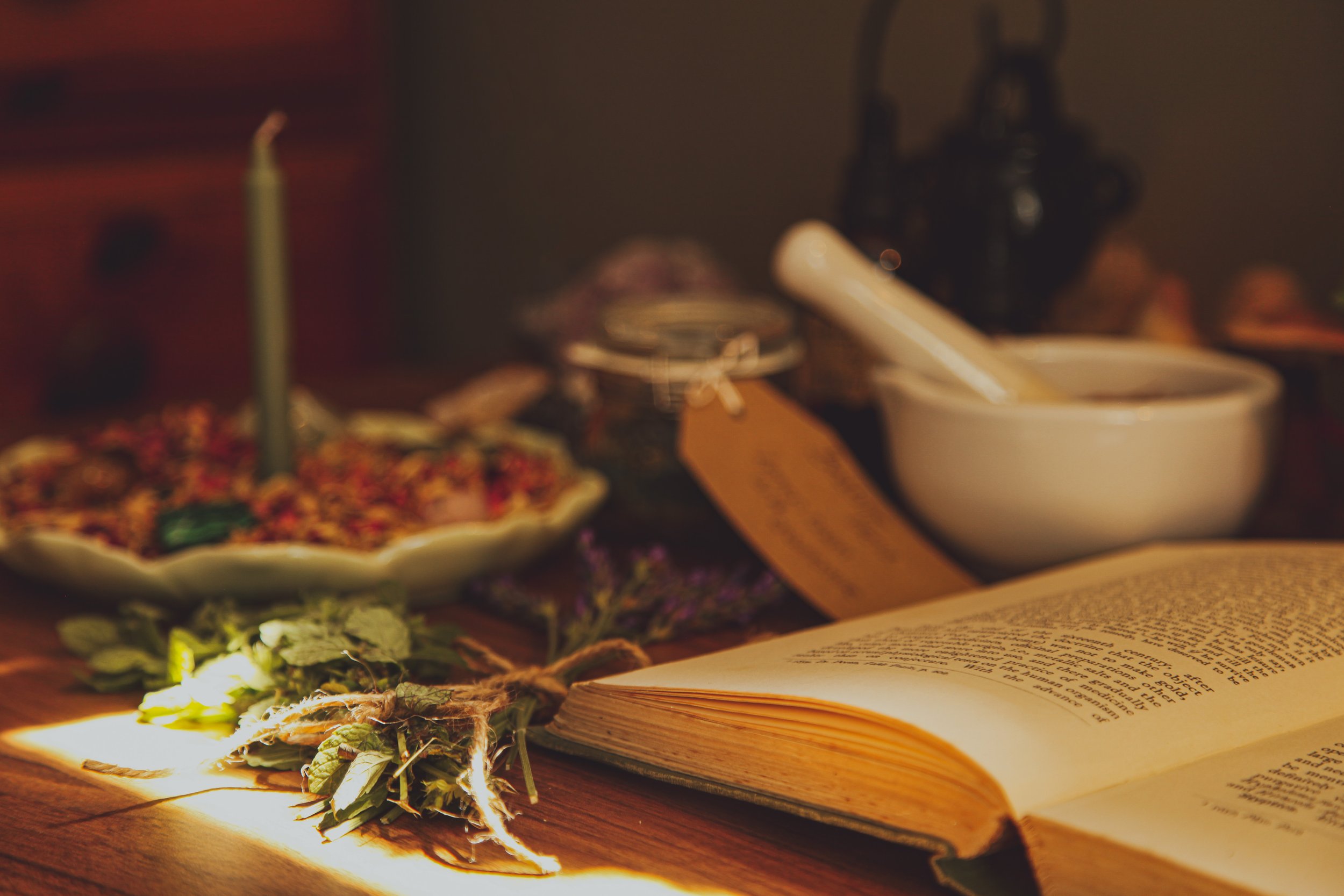 An open book with printed text is on a wooden table, illuminated by warm, soft lighting. Beside the book, there are dried leaves and a string of small lights. In the background, a ceramic dish with a candle, some jars, and blurred objects can be seen, creating a cozy, rustic atmosphere.