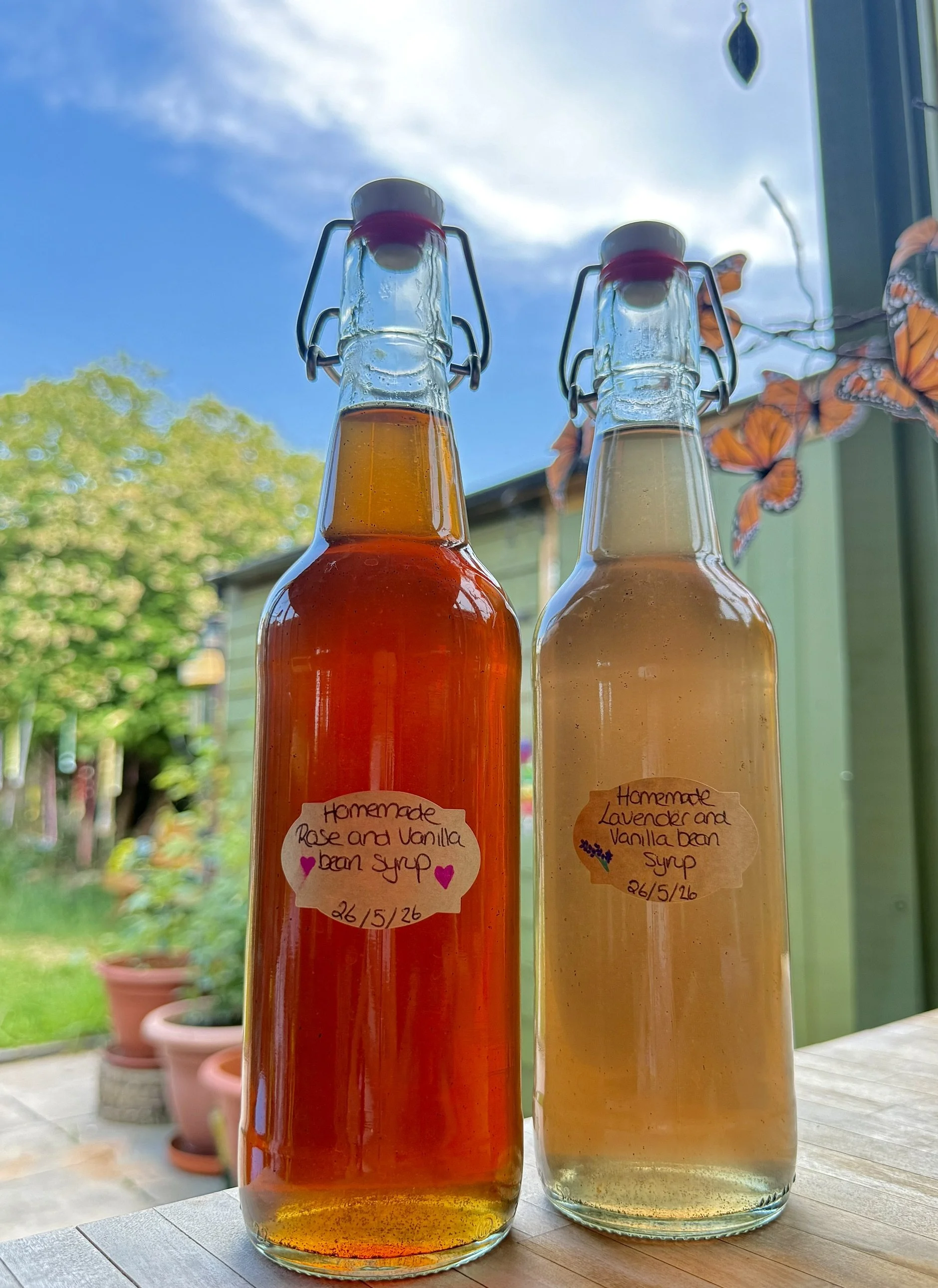 Two glass bottles filled with homemade flavored syrup, one with red syrup named "Rose and Vanilla Bean Syrup" and one with light yellow syrup named "Lavender and Vanilla Bean Syrup," both have flip-top stoppers. The bottles are on a wooden table near