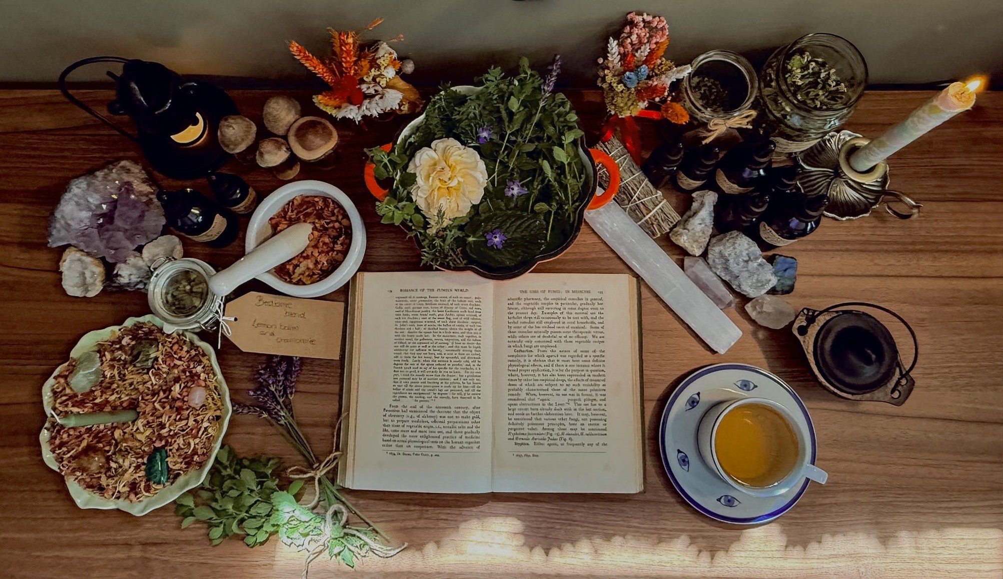 A top-down view of a wooden table displaying a spiritual or ritual setup. The setup includes an open book, several jars and containers of herbs and stones, a lit candle in a holder, a small cup of tea or herbal infusion, a dish with dried herbs, a potted plant with yellow flowers, various crystals, branches with leaves, and flowers. The arrangement appears to be used for meditation, herbalism, or spiritual practices.