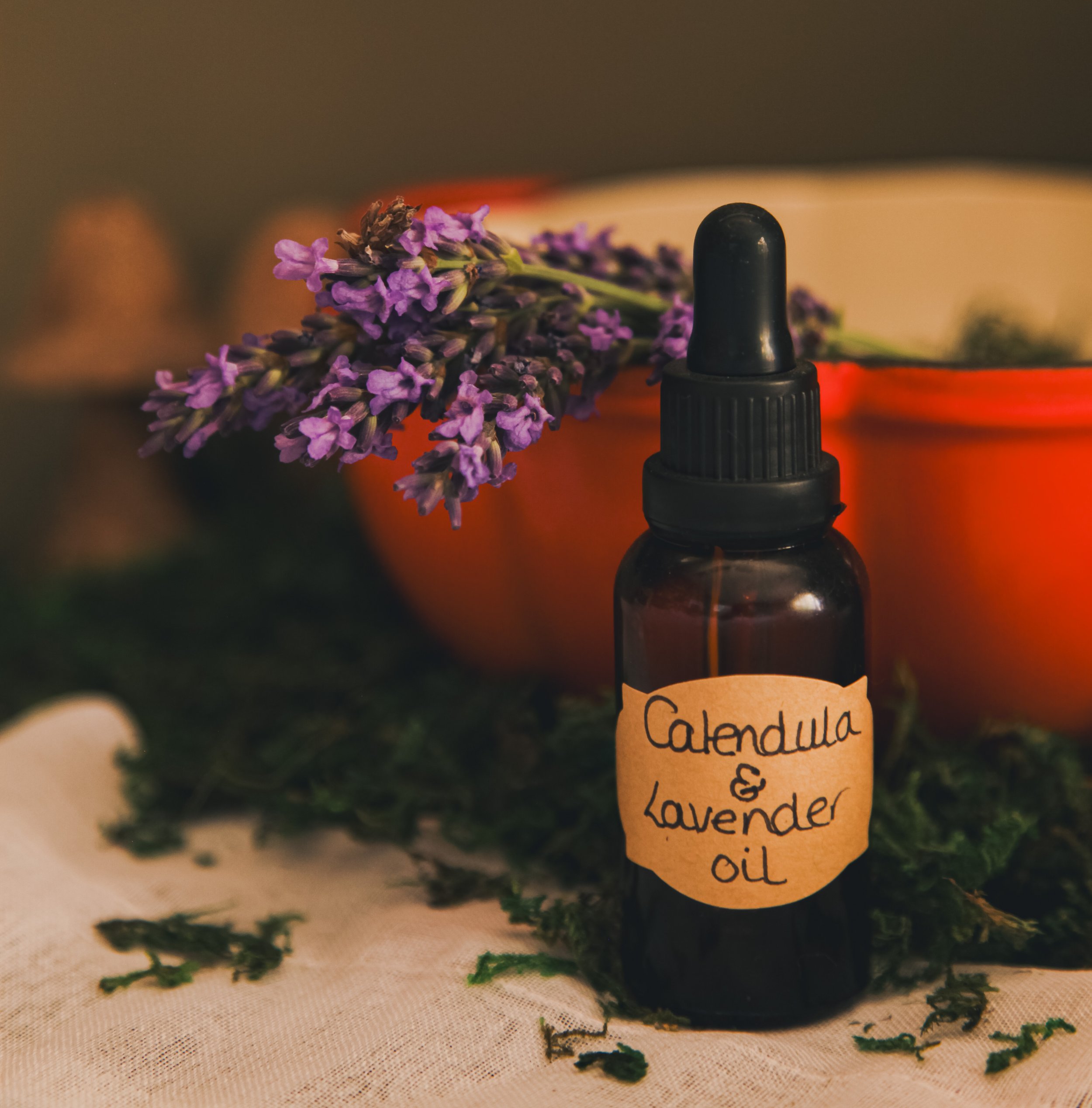 A small brown glass bottle with a black dropper cap, labeled 'Calendula & Lavender Oil,' placed on a surface with dried lavender flowers and scattered green leaves around it.