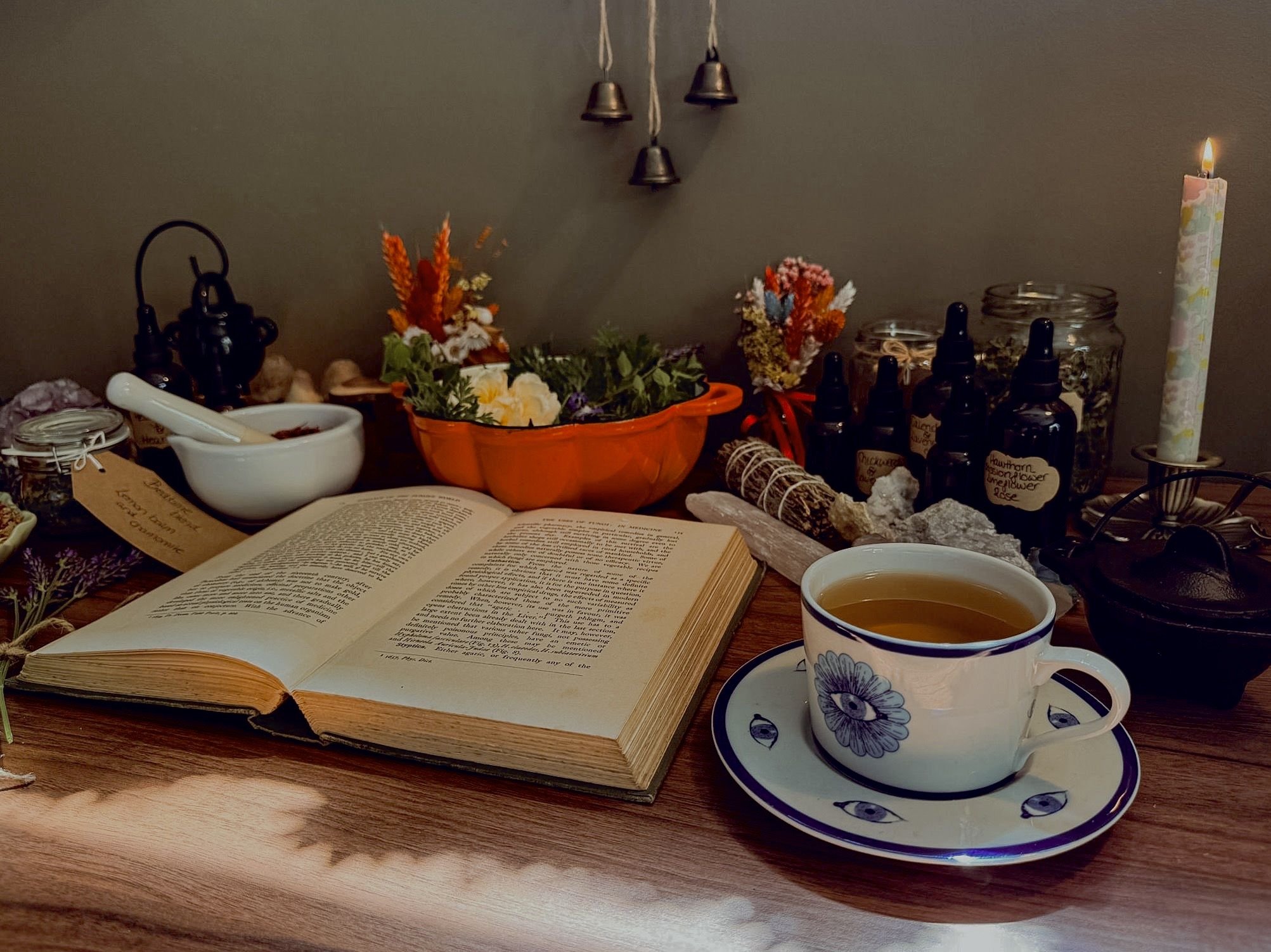 An open book, a cup of tea in a floral cup and saucer, and a variety of small bottles with dropper caps are arranged on a wooden table. Behind them, there are colorful dried flowers, small bells hanging on the wall, and a lit candle in a floral holder, all creating a cozy, bohemian atmosphere.