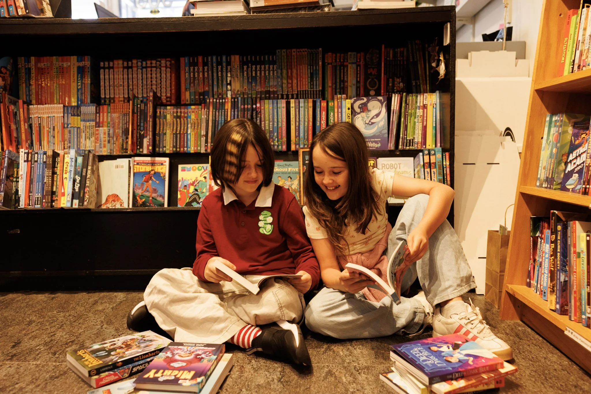Two children sitting on the floor in a bookstore, reading comic books, with shelves of books behind them.