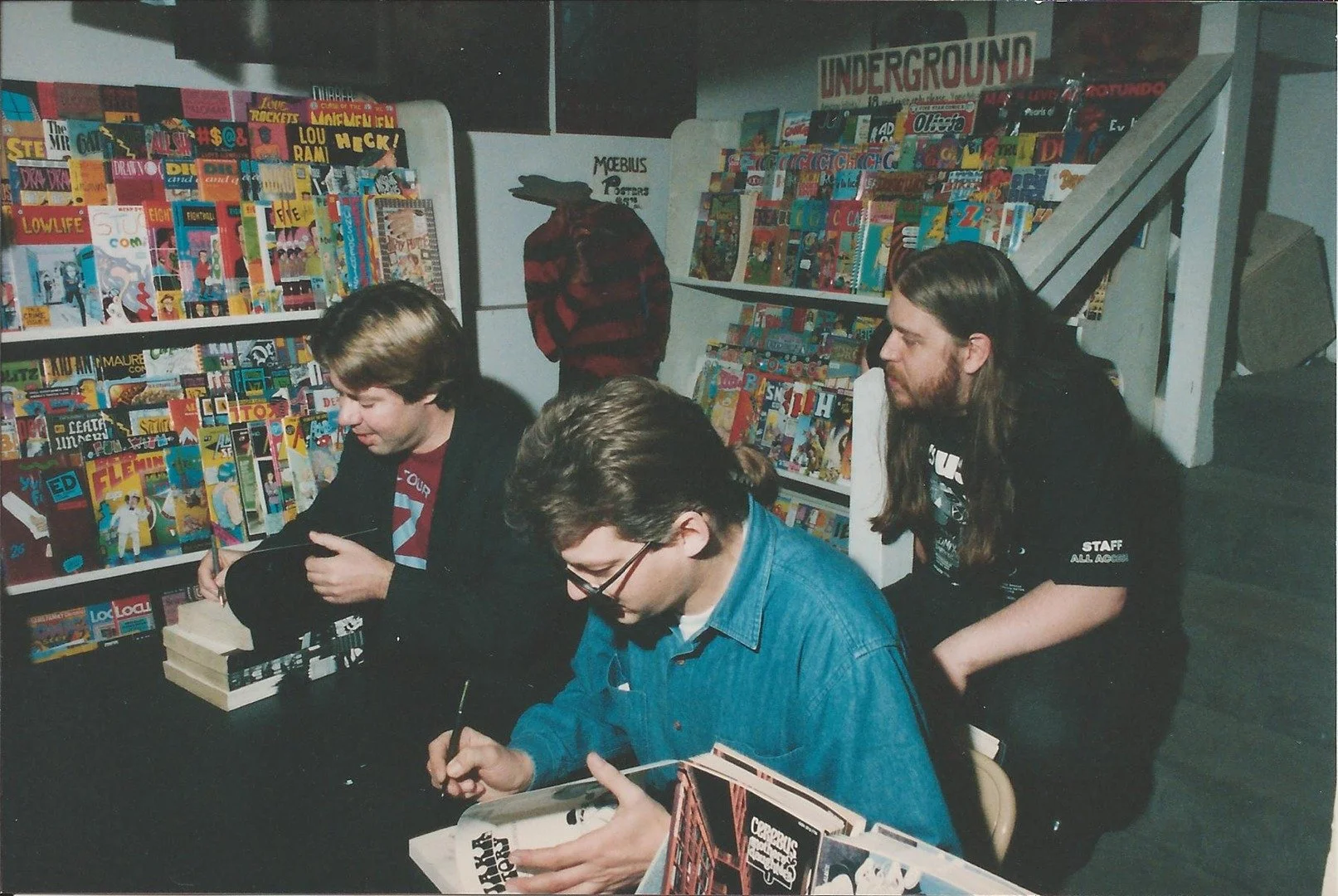 Three people sitting at a table, signing books at a comic book event. A fourth person in the background stands near a magazine rack.