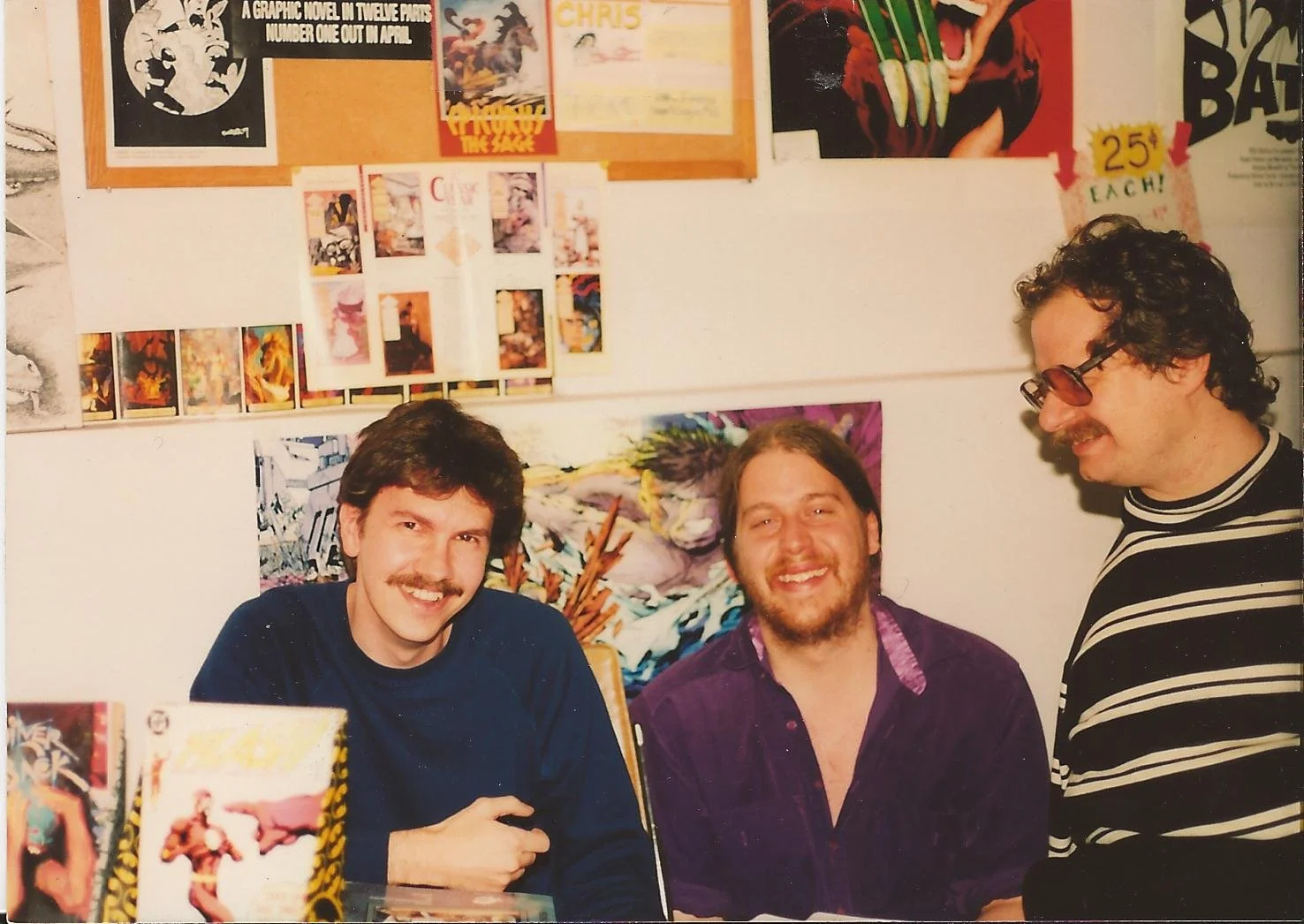 Three men sitting at a table surrounded by comic book posters and artwork on a wall.