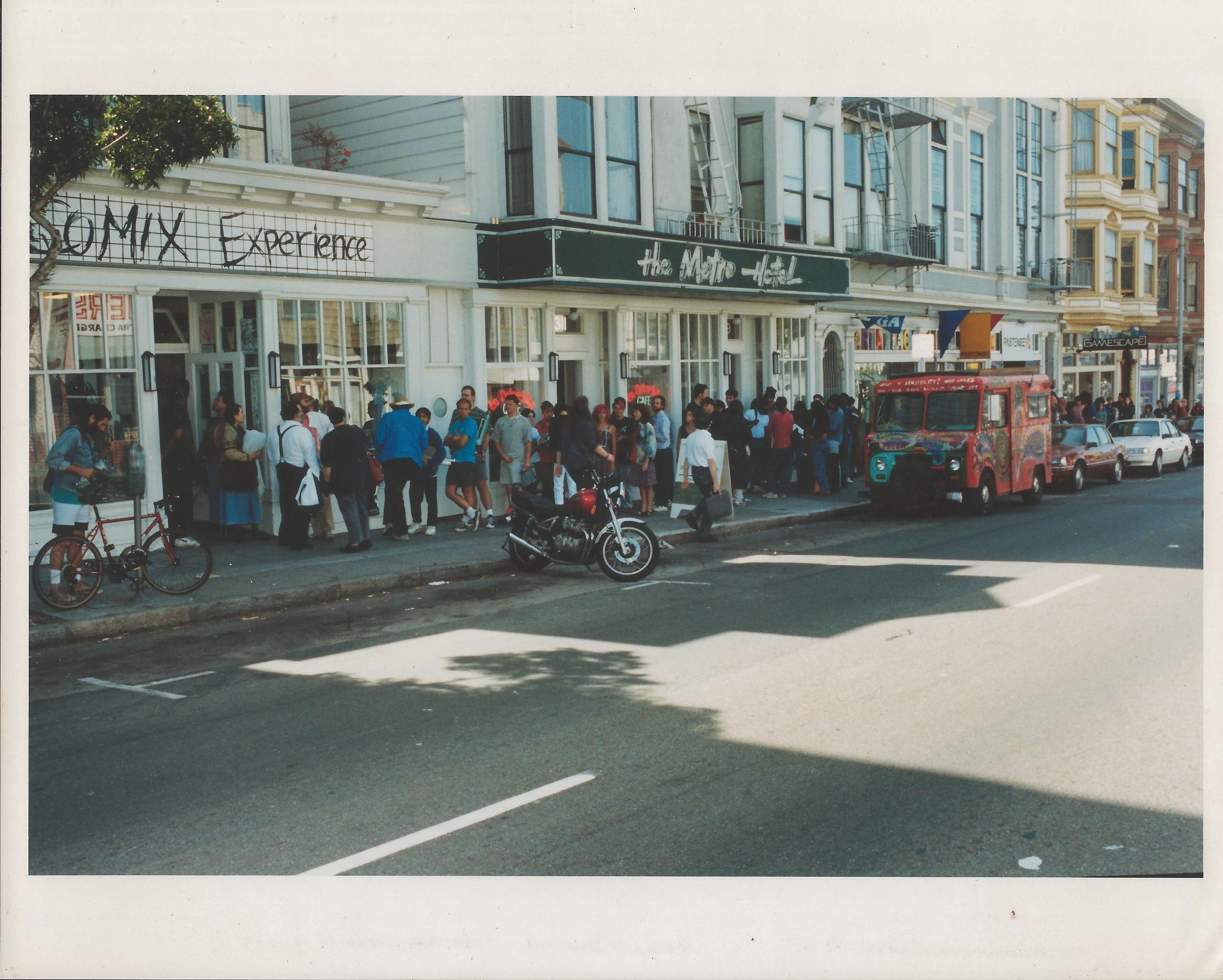 Street scene with a crowd of people gathered outside The Metro Hotel, a colorful van, and neighboring buildings with signage and apartments.