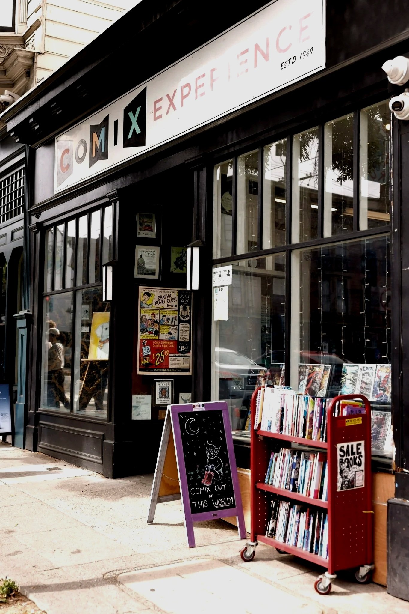 Outside of a comic bookstore with books for sale on a red cart and a chalkboard with a drawing of a cat and moon.