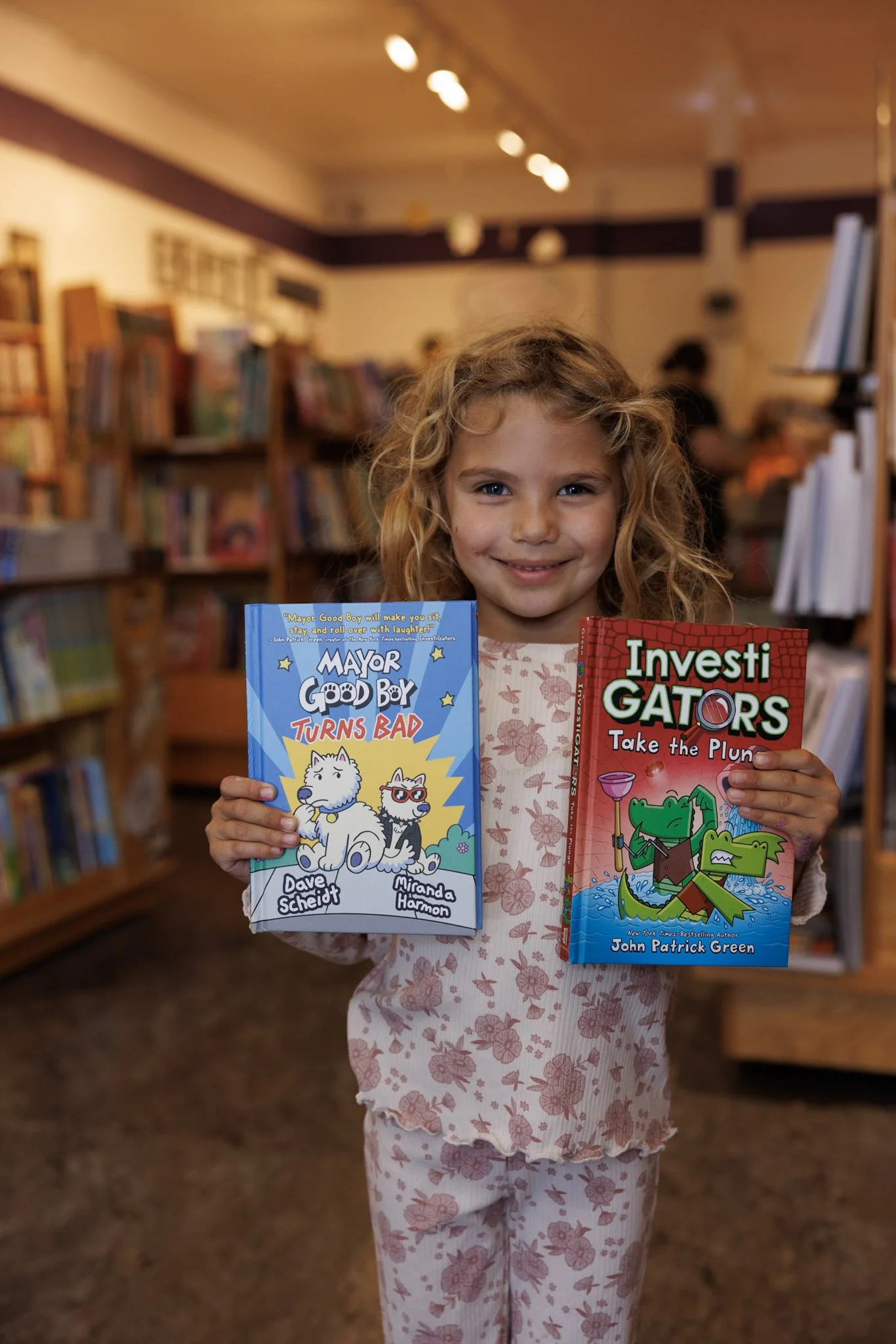 A young girl with curly hair smiling in a bookstore, holding two children's books titled 'Mayor Good Boy Turns Bad' and 'Investi Gators Take the Plun', with bookshelves in the background.