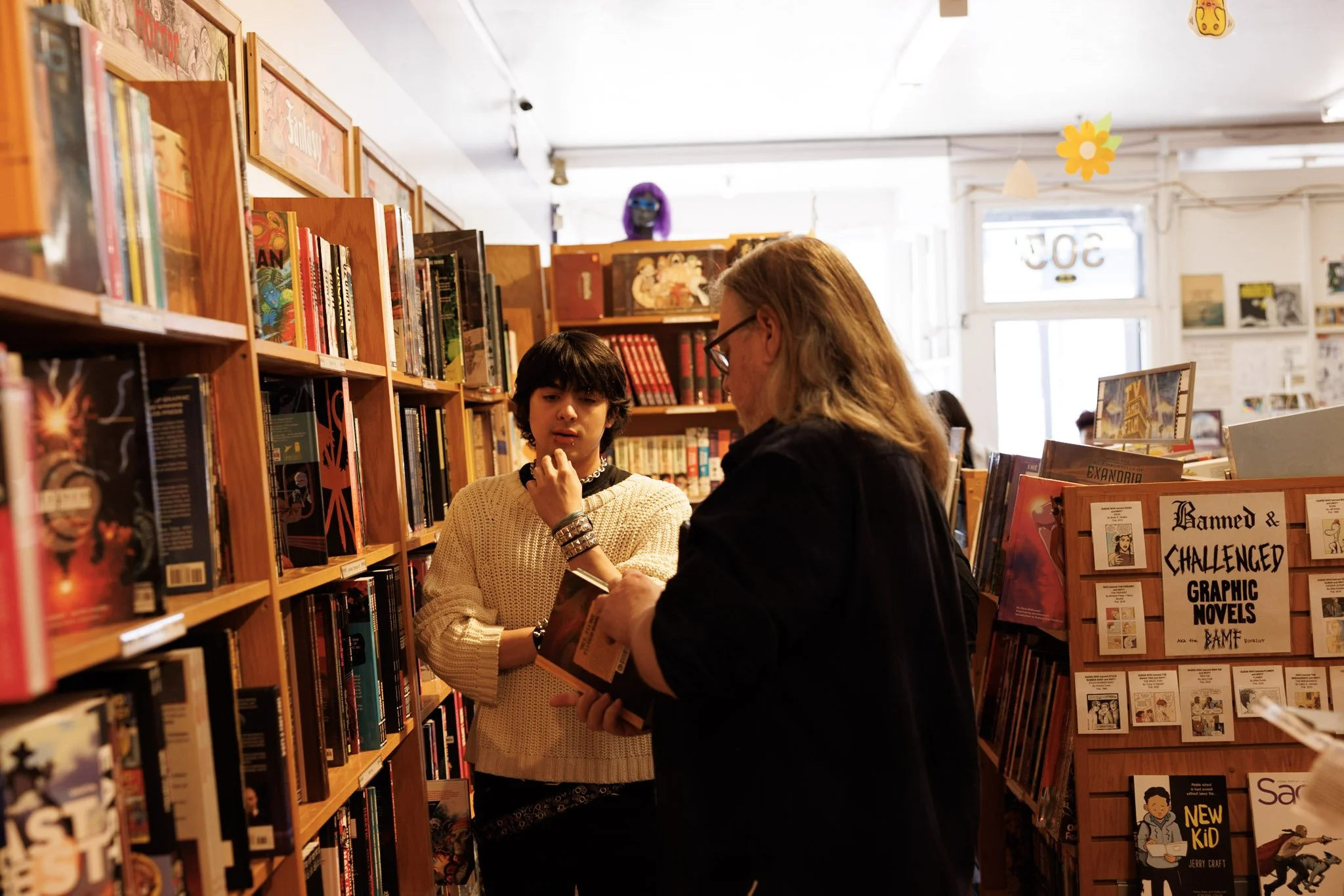 Two women shopping for graphic novels in a bookstore, with wooden shelves filled with books around them. One woman has light brown hair and glasses, the other has dark hair and is wearing a beige sweater. They are looking at a book together. Hanging decorations and more books are visible in the background.