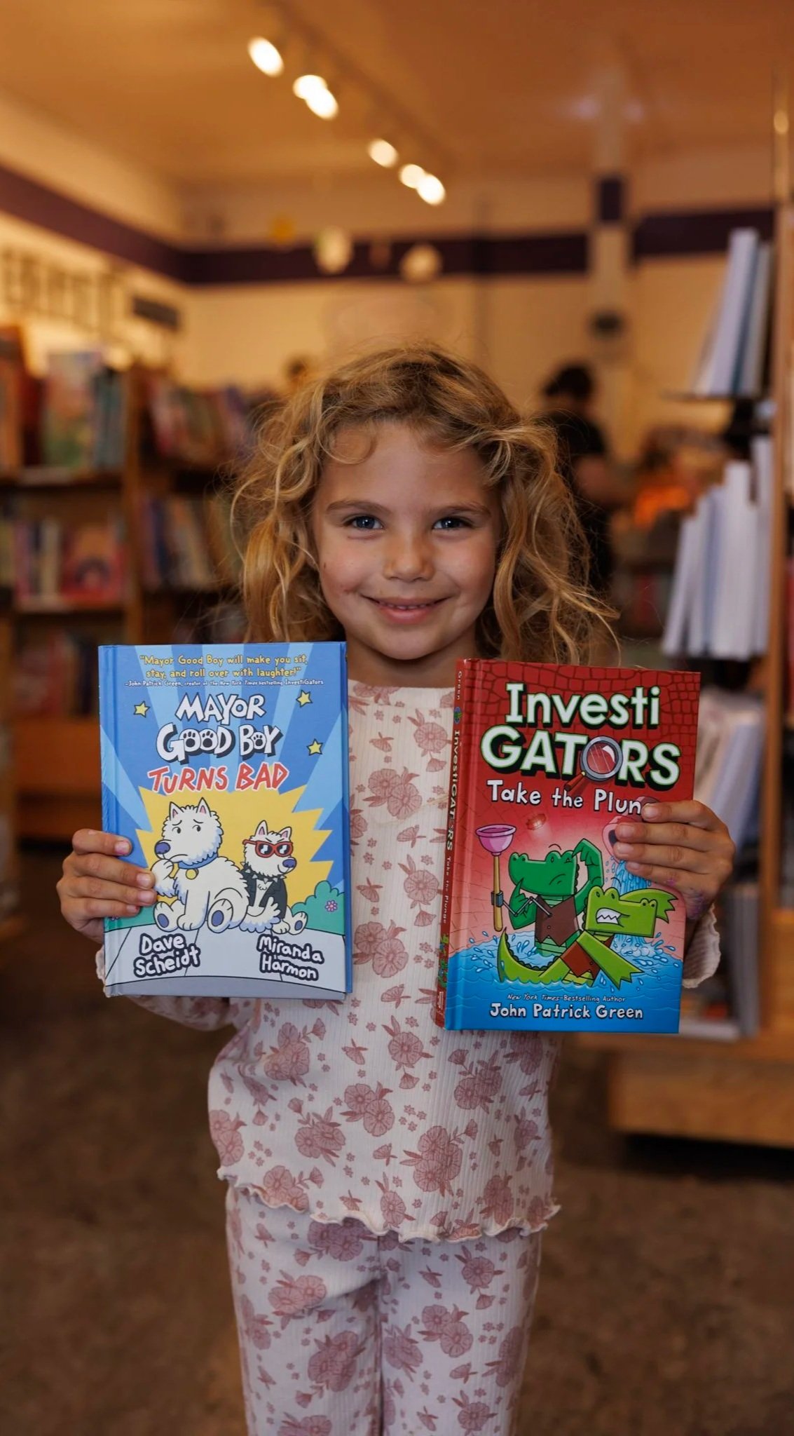 A young girl with curly hair smiling and holding two children's books, one titled 'Mayor Good Boy Turns Bad' with a white dog and a black and white dog on the cover, and the other titled 'InvestiGATORS Take the Plunge' with an illustration of crocodile characters in a boat, inside a bookstore.