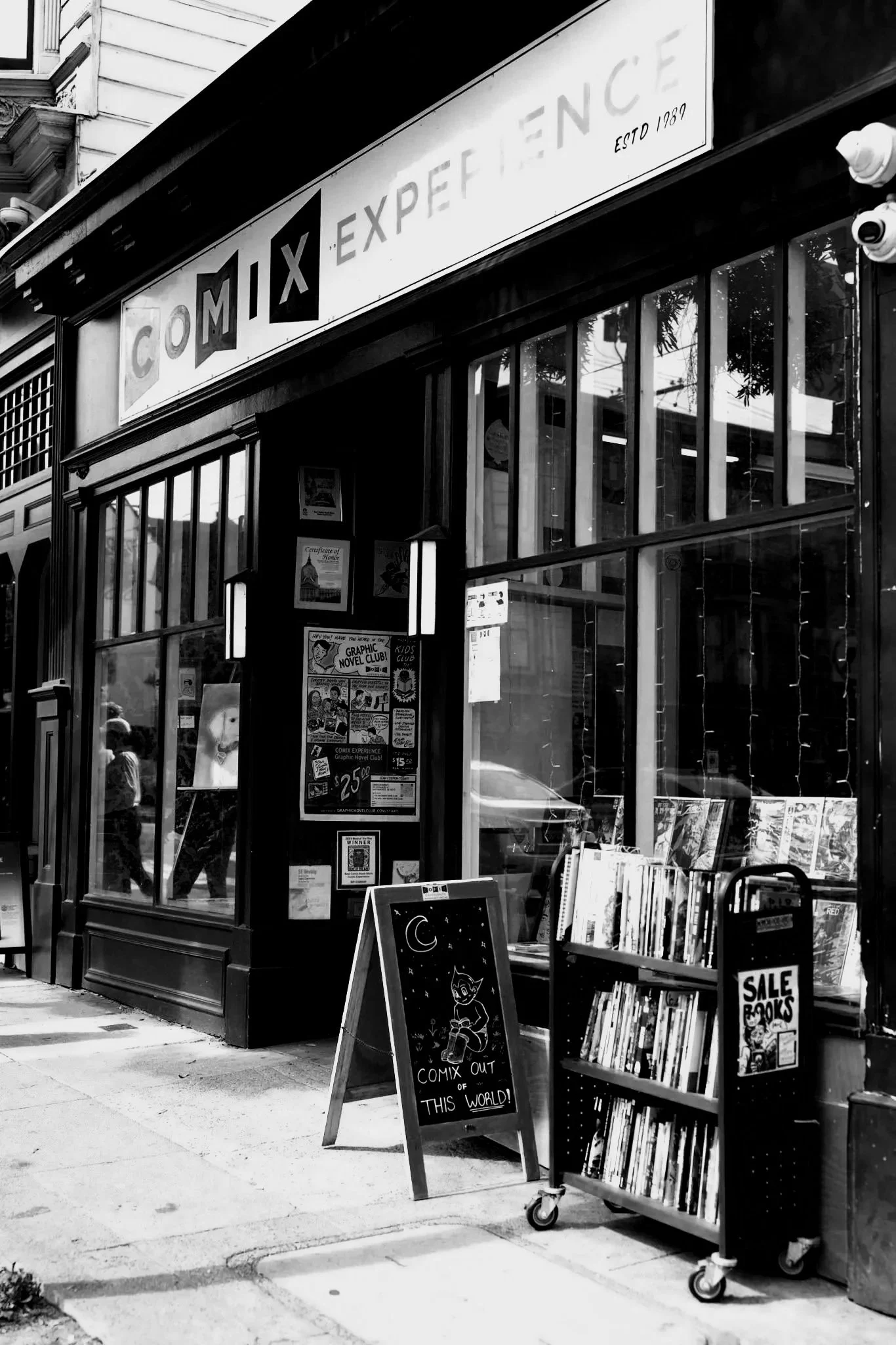 A bookstore storefront with a sign that reads 'COMIX EXPERIENCE' and 'ESTD 1989'. There are books on display outside, including a sign that says 'SALE BOOKS' and a chalkboard sign with a cartoon cat and the phrase 'COMIX OUT OF THIS WORLD!' The window shows reflections of patches of sky and tree leaves.