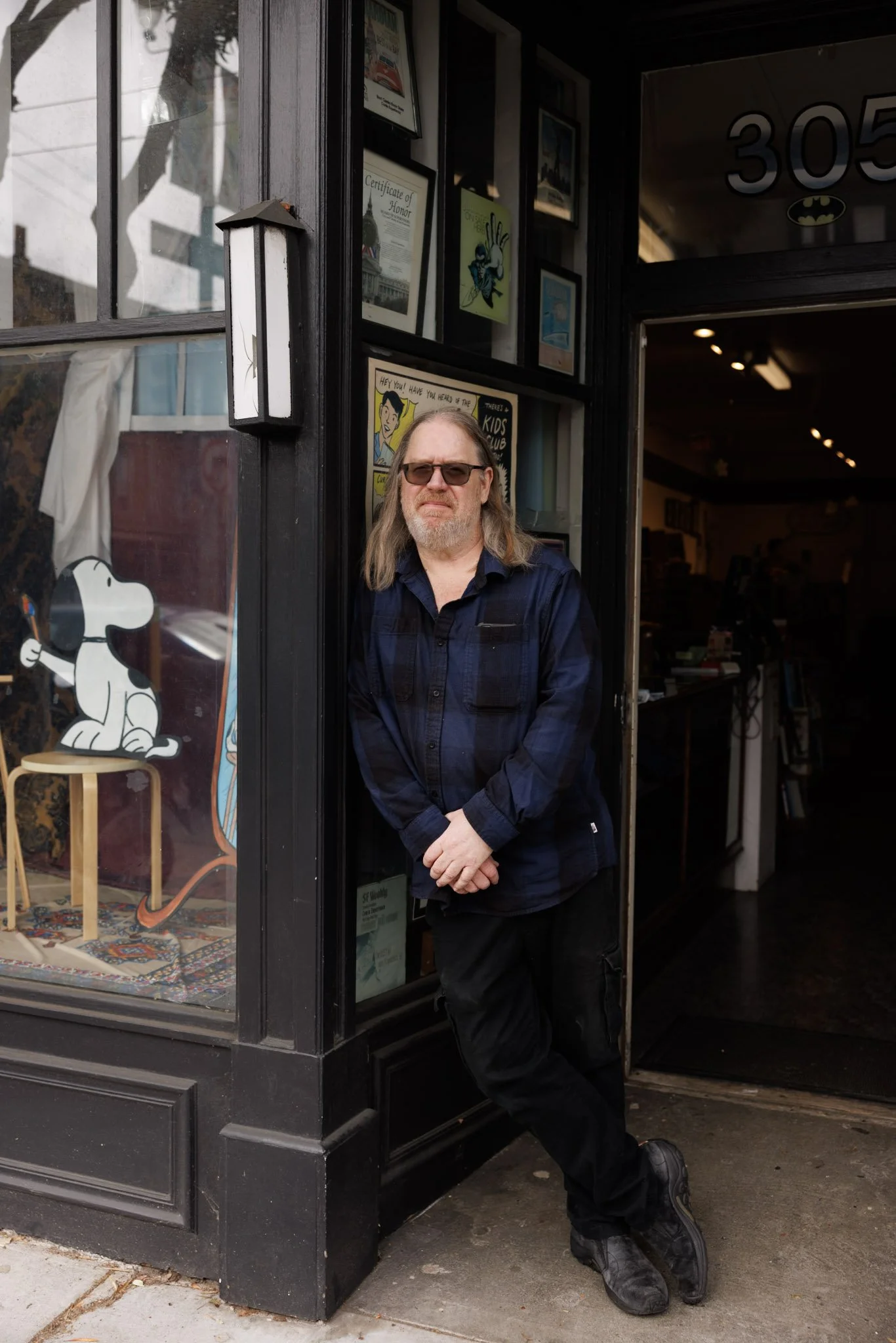 A man with long hair, a beard, and sunglasses wearing a dark blue shirt and black pants is standing in an open doorway of a shop. The shop has black framed windows with colorful posters and awards displayed inside. A cartoon dog figure is visible through the window on the left.