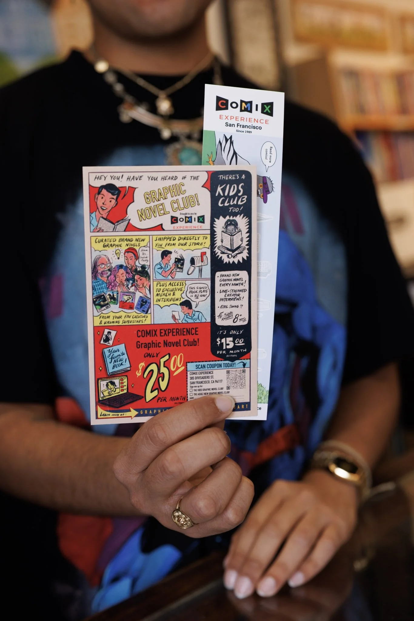 Person holding a colorful flyer advertising a comic experience and graphic novel club, with bookshelves in the background.